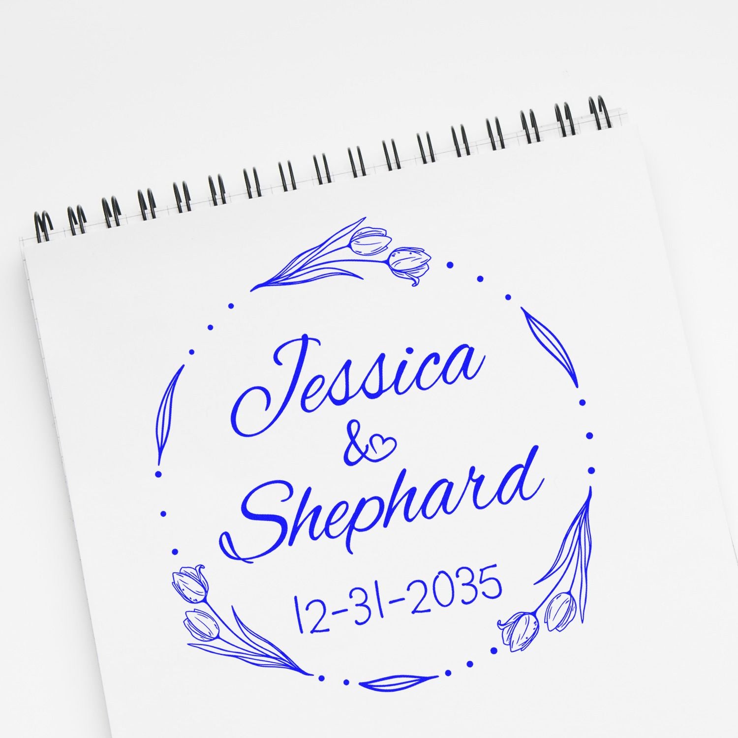 Blue custom wedding stamp and seal on white spiral notebook, reading Jessica & Shephard with floral wreath and 12-31-2035.