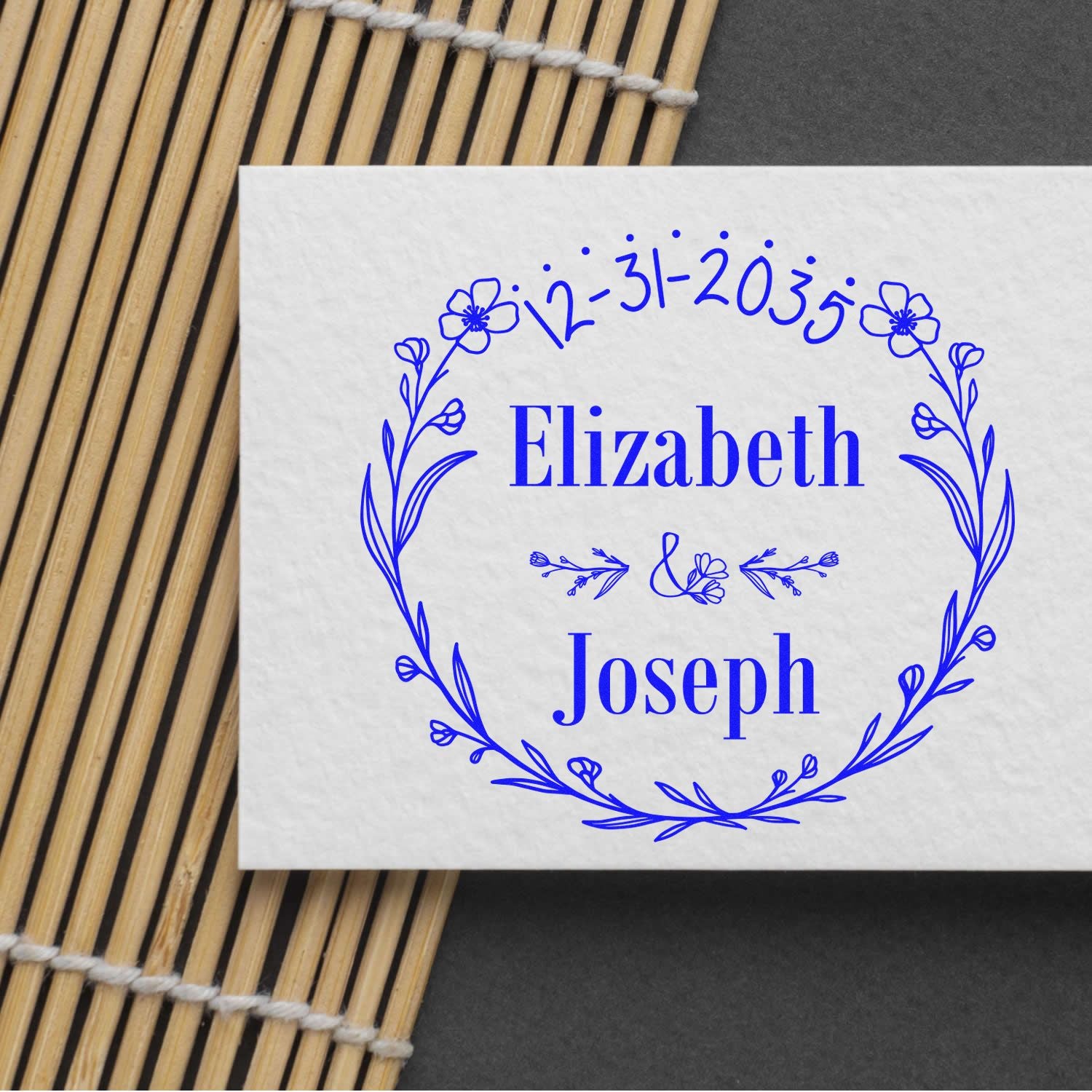 Blue custom stamp and seal with floral wreath, Elizabeth and Joseph, 12-31-2035, on white paper and bamboo mat.
