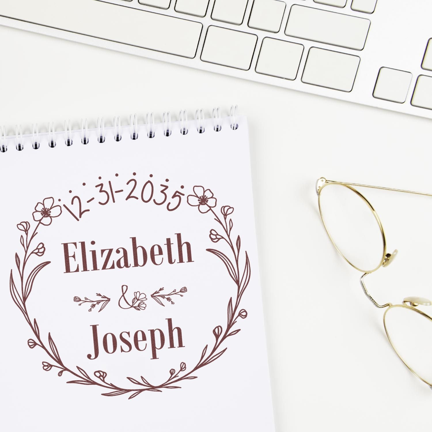 Brown custom floral stamp seal for Elizabeth and Joseph with date on white notebook, beside gold eyeglasses and keyboard.