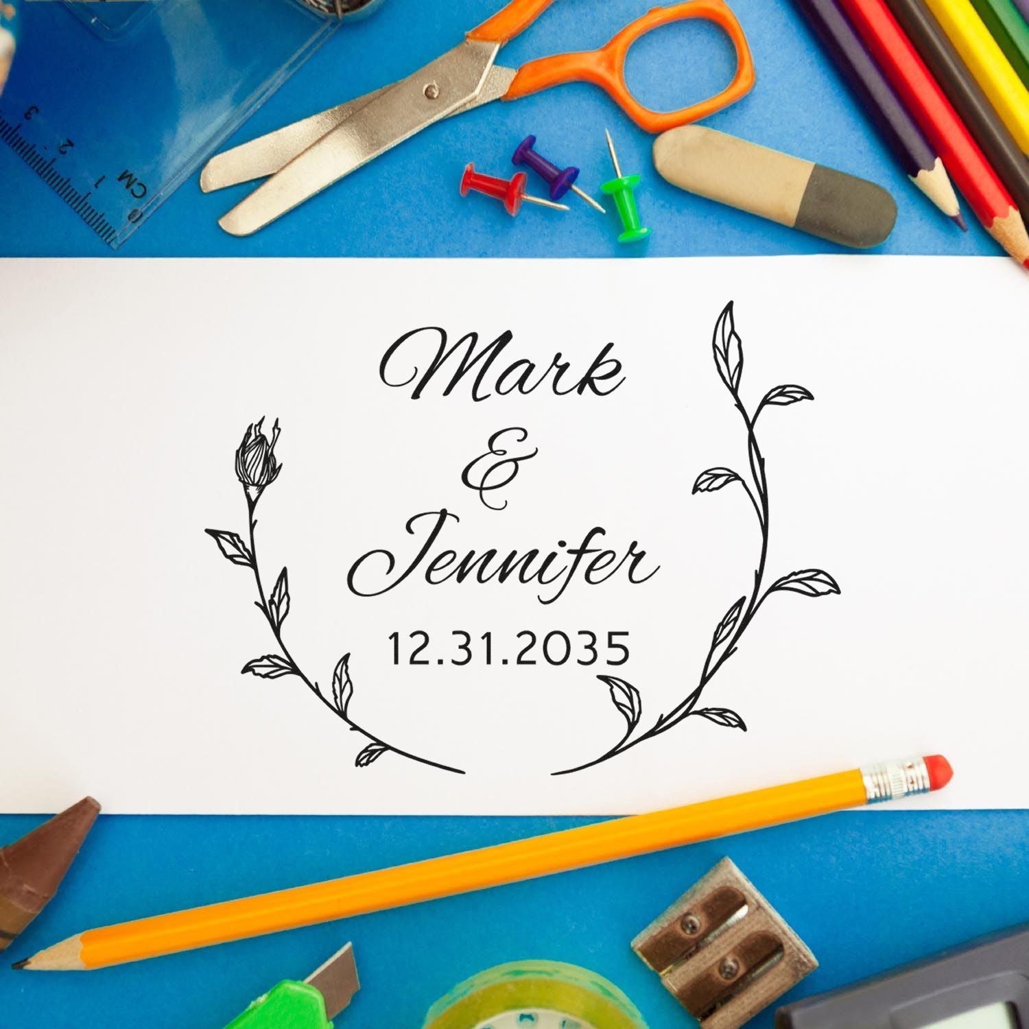 Custom stamp and seal reading Mark & Jennifer 12.31.2035, leafy wreath on white paper with blue background and craft tools.
