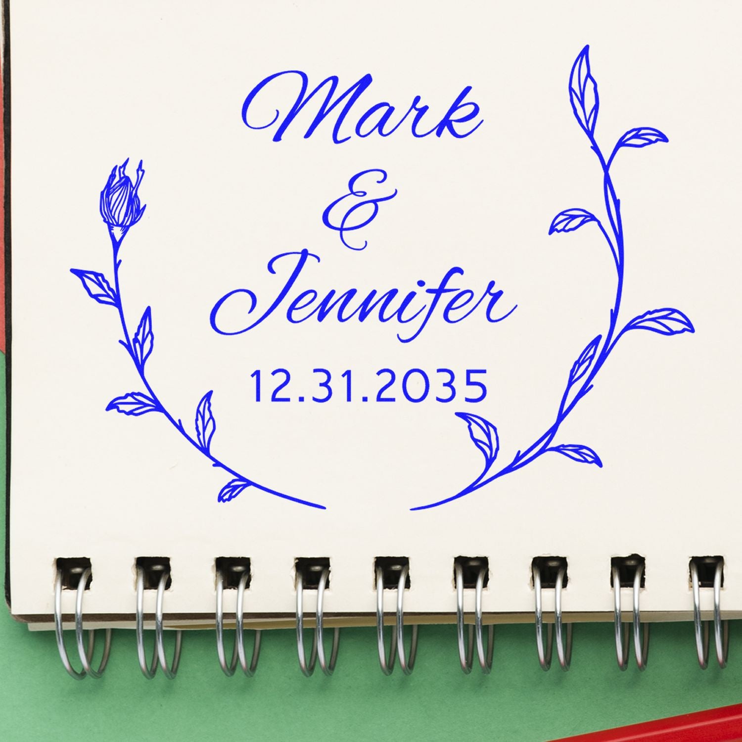 Blue custom wedding stamp seal on cream notebook page with floral wreath, reading Mark & Jennifer and 12.31.2035.