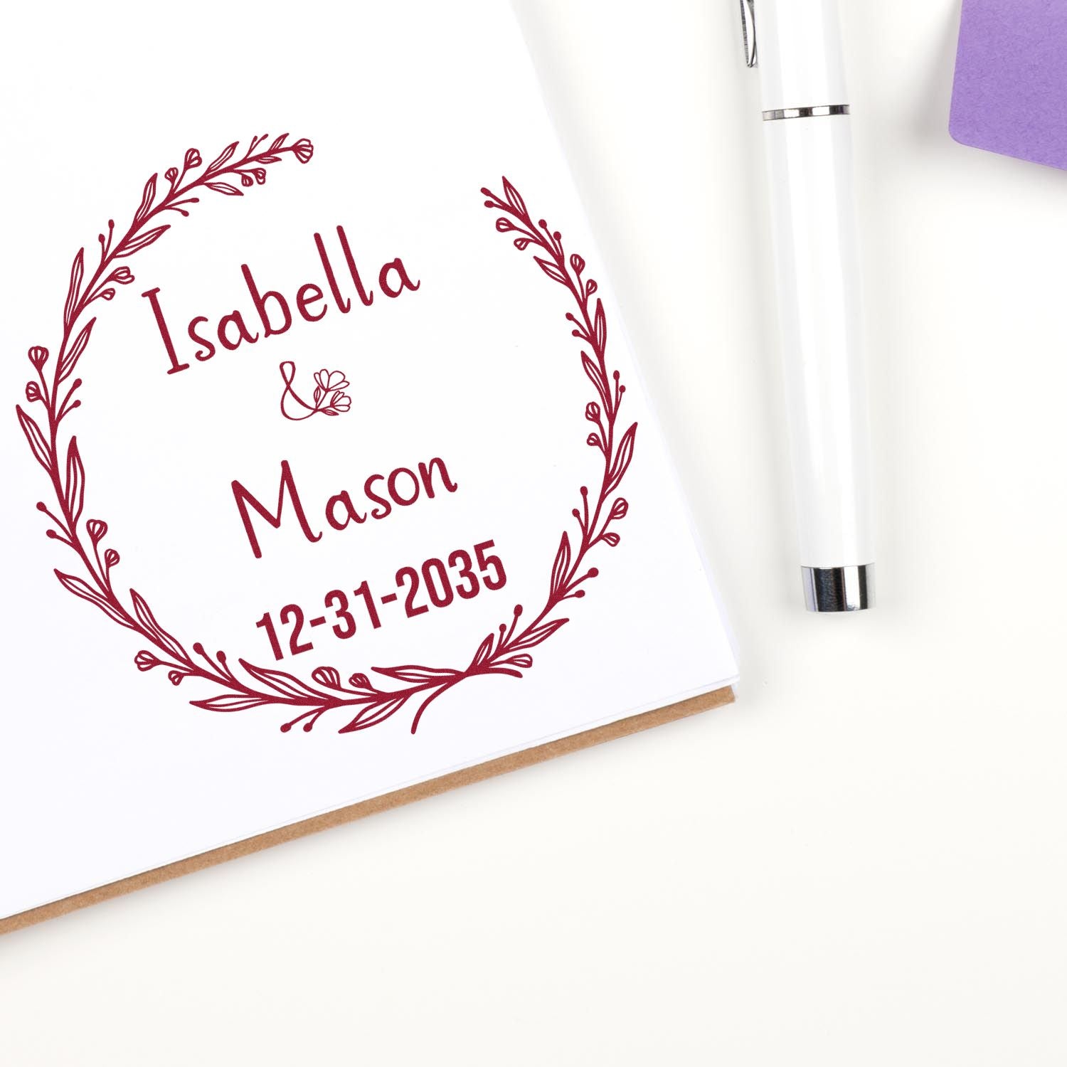 Burgundy floral wreath stamp with Isabella Mason and 12-31-2035, beside a white seal pen on white paper