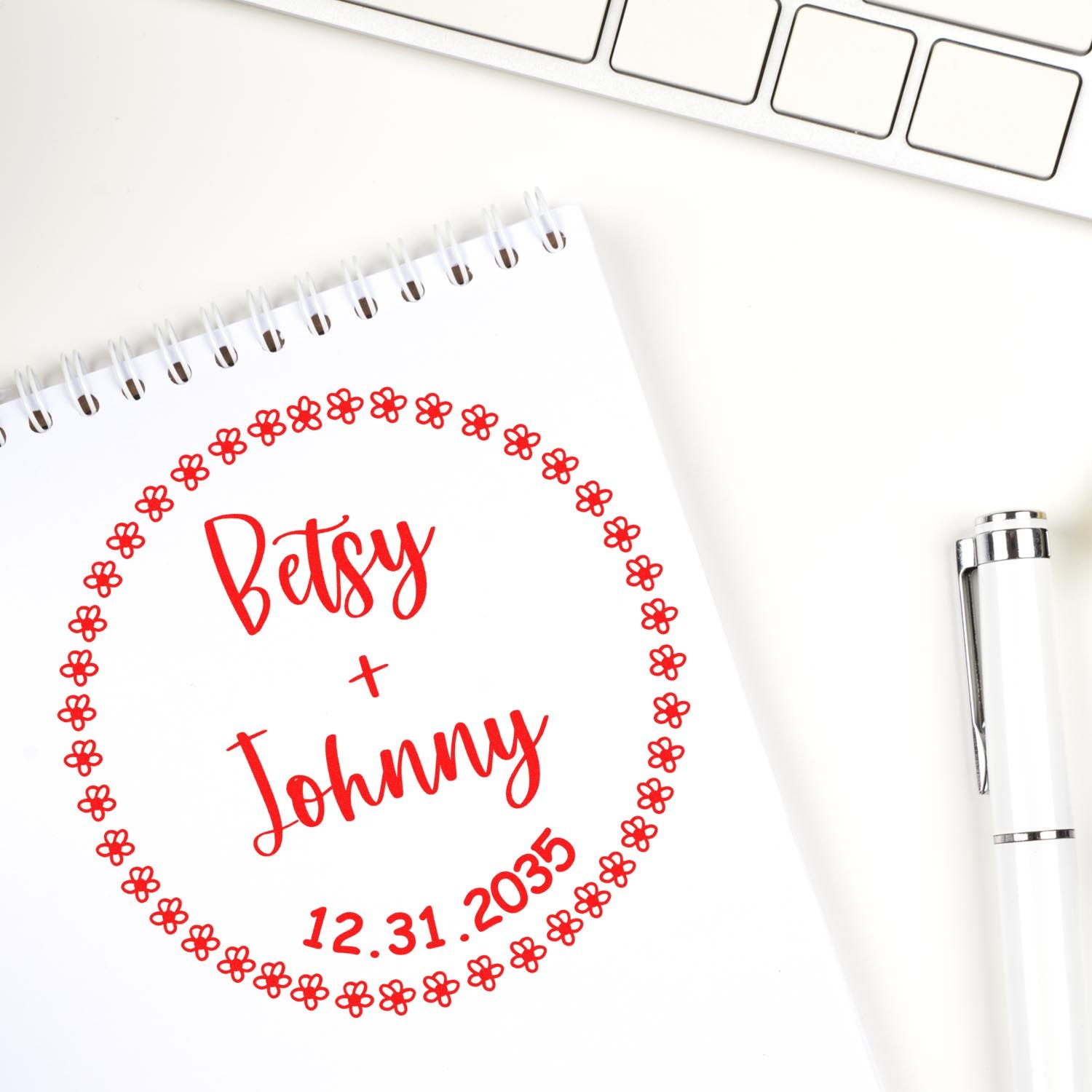 Custom red floral wedding stamp seal with Betsy + Johnny and 12.31.2035 on a white spiral notebook beside a silver pen.