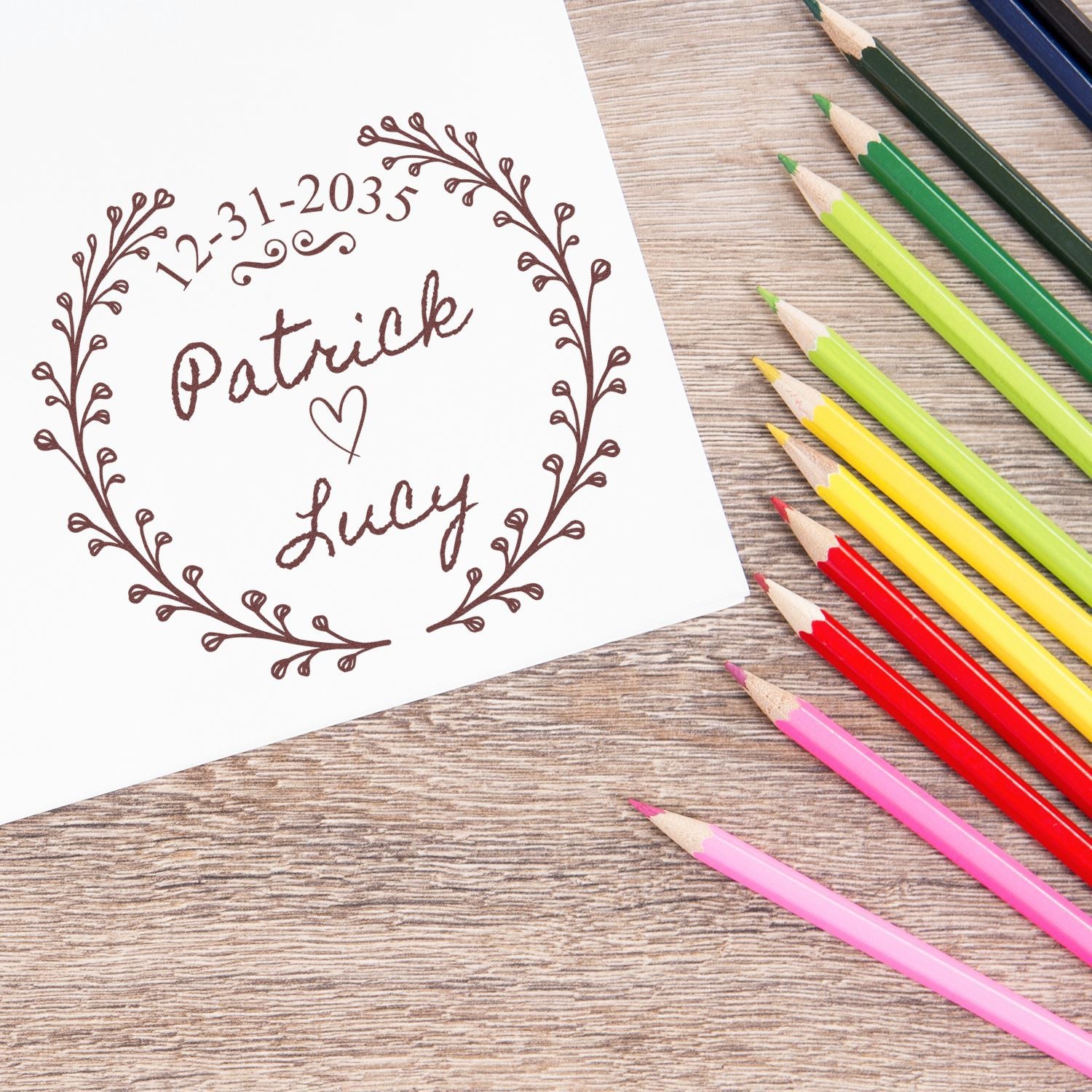 Custom brown stamp seal reading Patrick and Lucy inside a wreath on white paper, with colorful pencils on wood