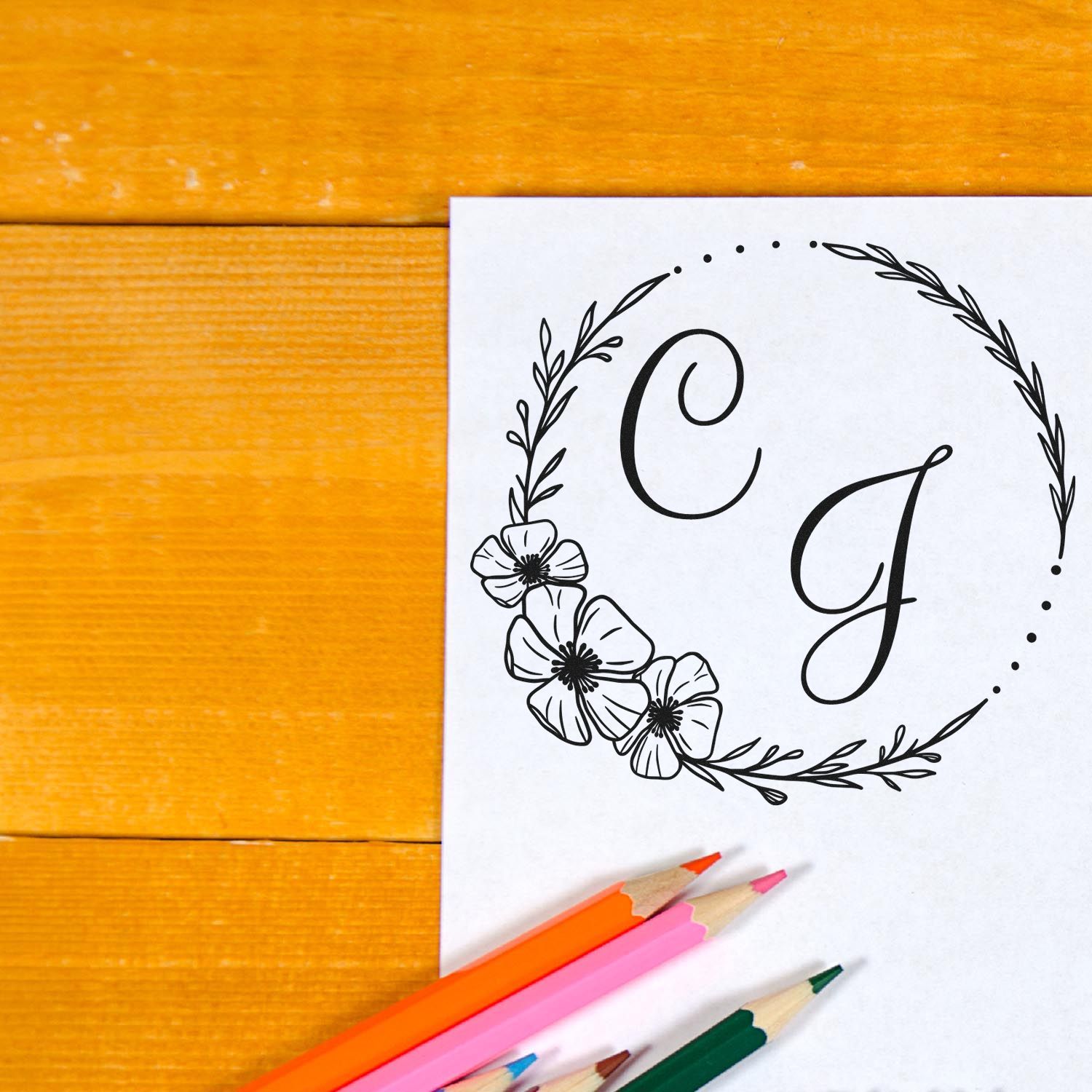 Black floral monogram stamp and seal design with letters C and J on white paper, beside colored pencils on a warm orange wood table.