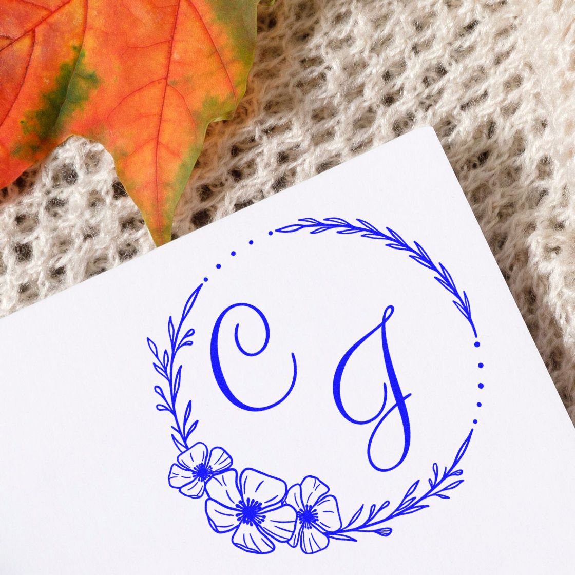 Blue custom stamp seal with initials CJ in a floral wreath on white paper, beside an orange autumn leaf on beige knit.