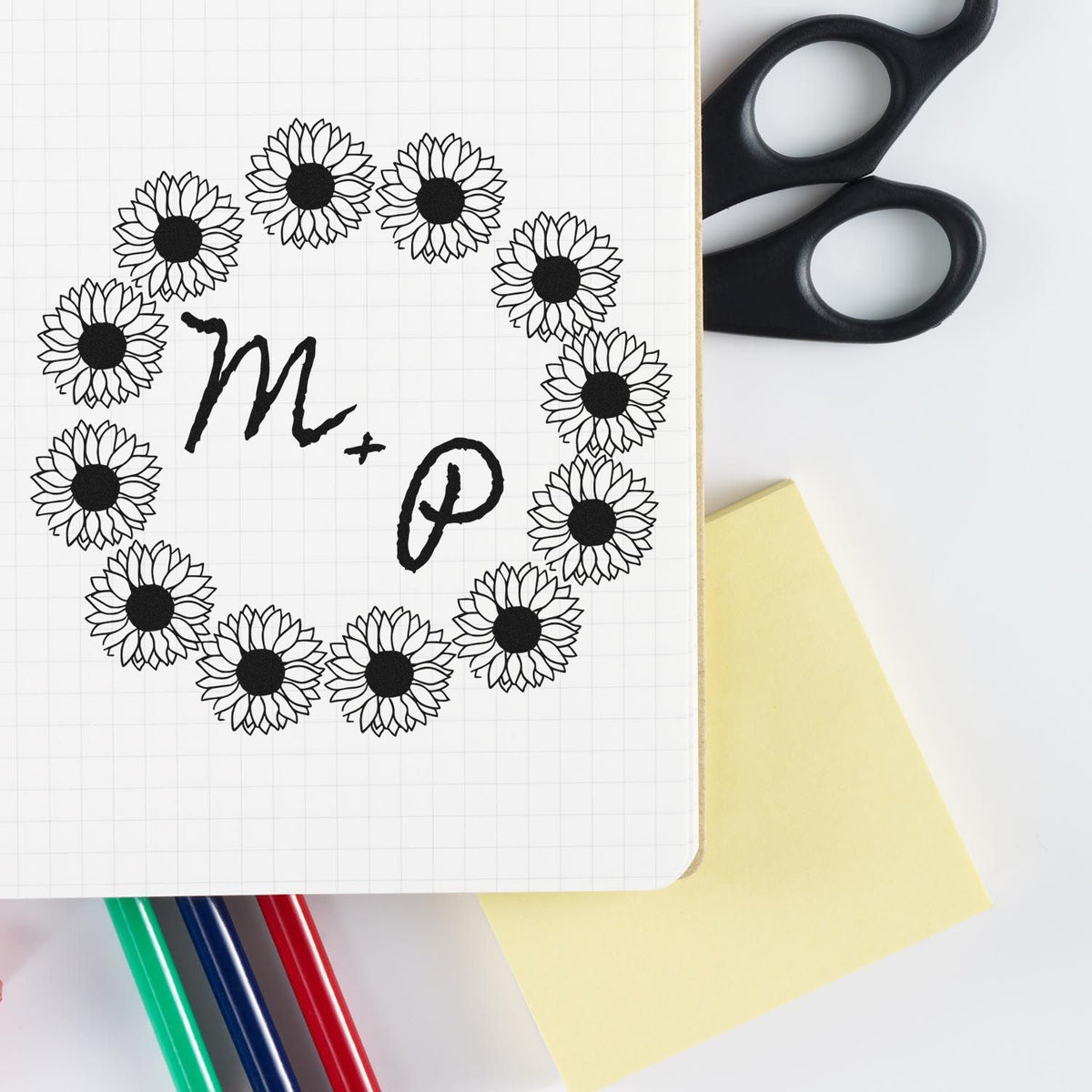 Black sunflower stamp and seal around M+P monogram on grid paper, with black scissors, yellow note, and colored pens.