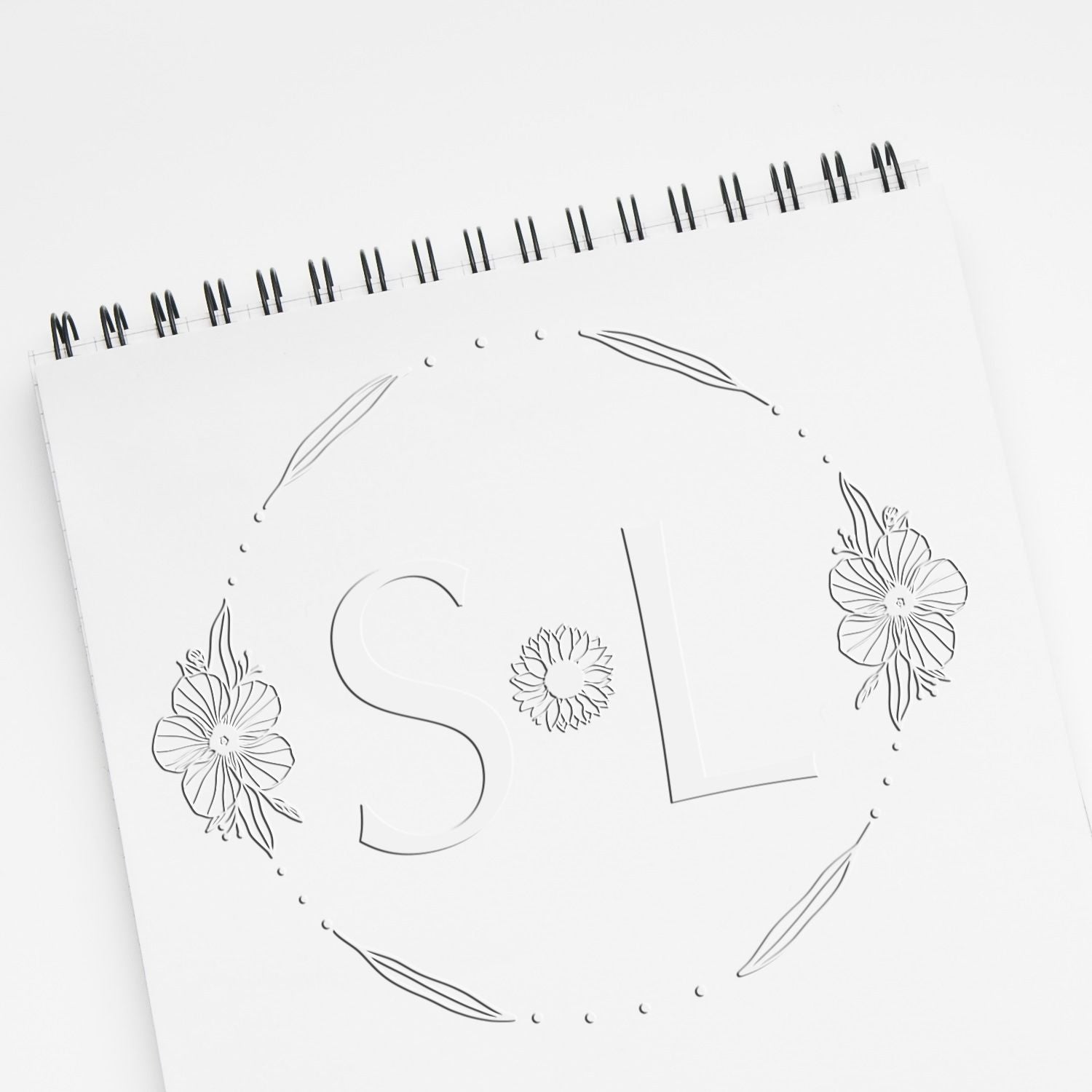 White spiral notebook with embossed stamp and seal design, black floral line art, and subtle gray monogram letters.