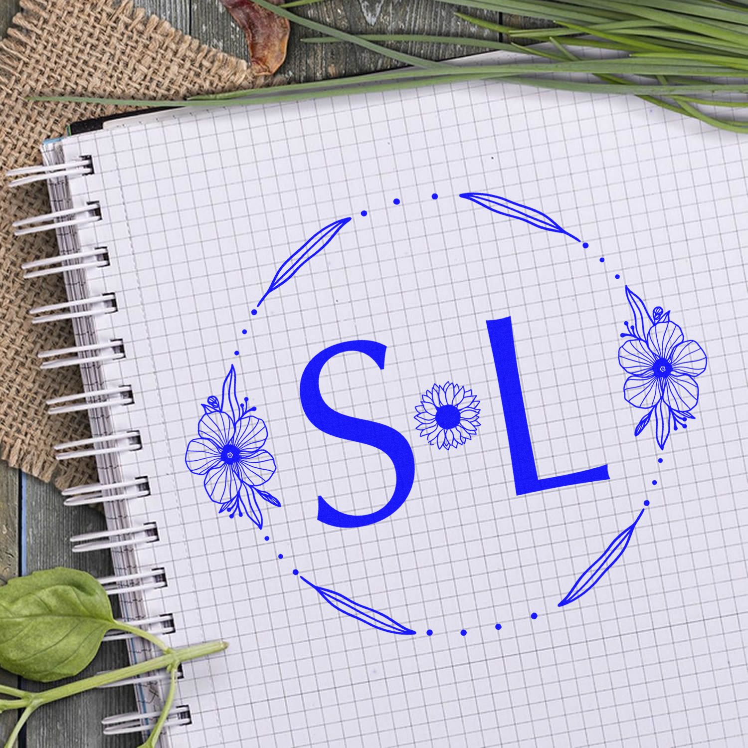 Blue SL stamp seal logo with floral wreath on grid notebook, surrounded by green leaves