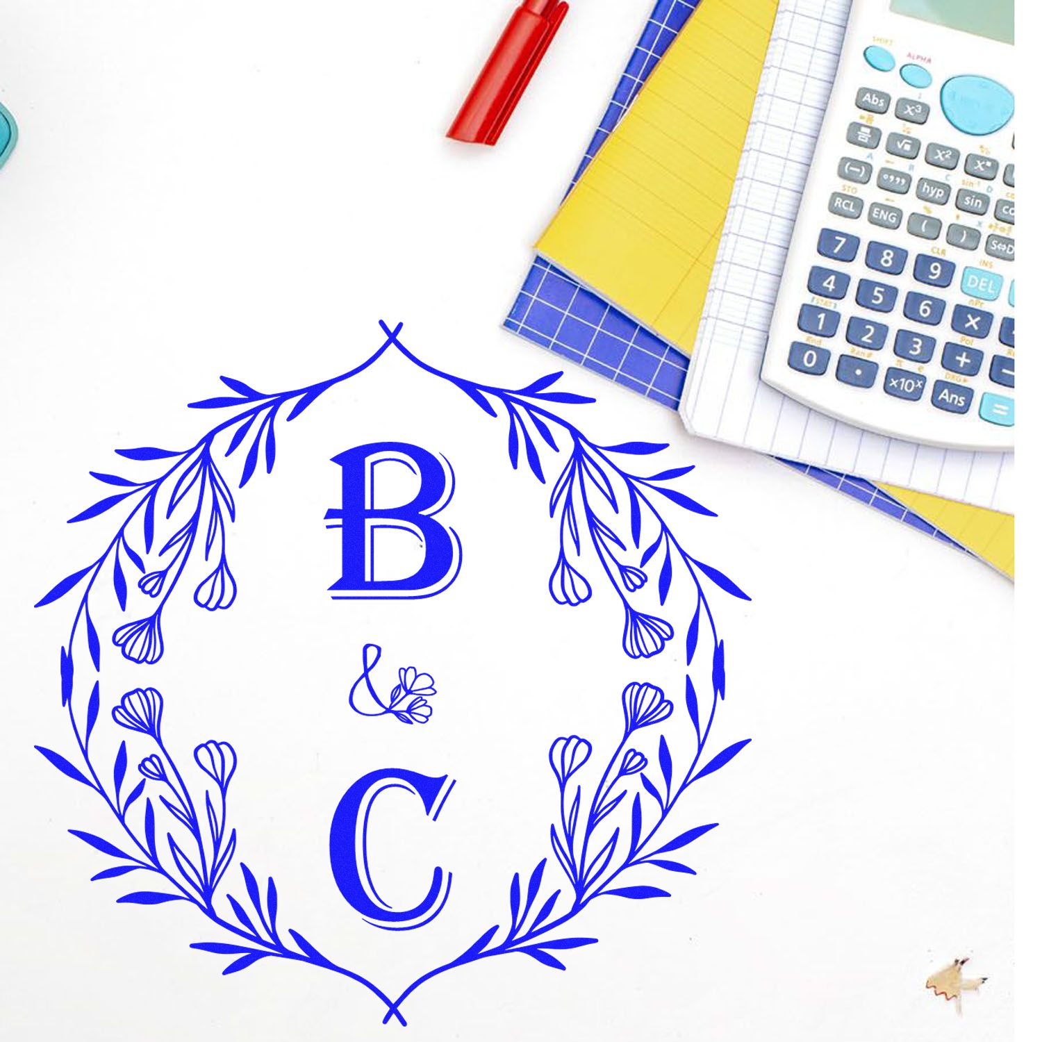 Blue floral B&C custom stamp seal with wreath monogram on white, beside colorful notebooks and a calculator.