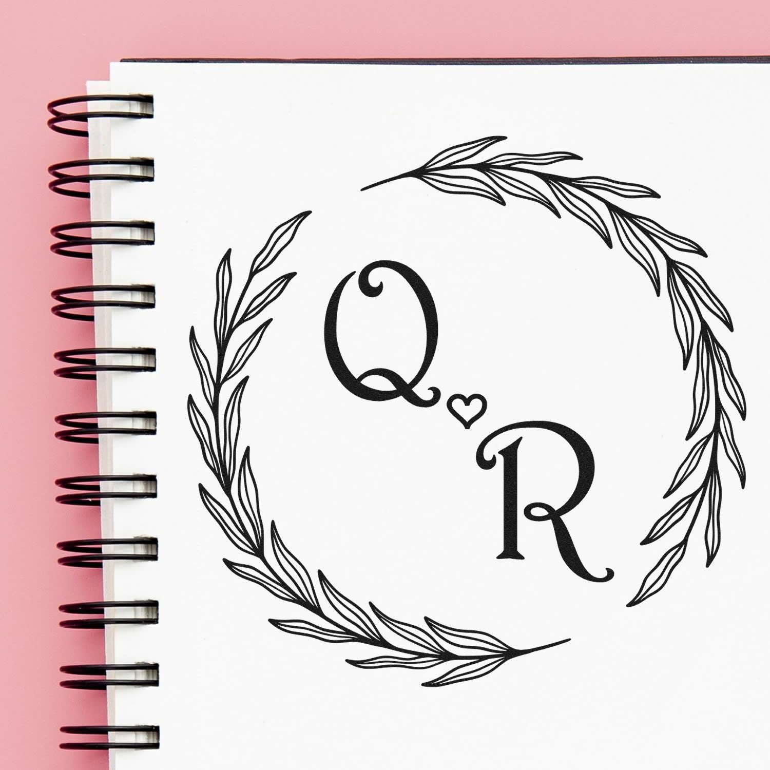 Black Q heart R monogram stamp and seal with botanical wreath on a white spiral notebook, against a pink background.