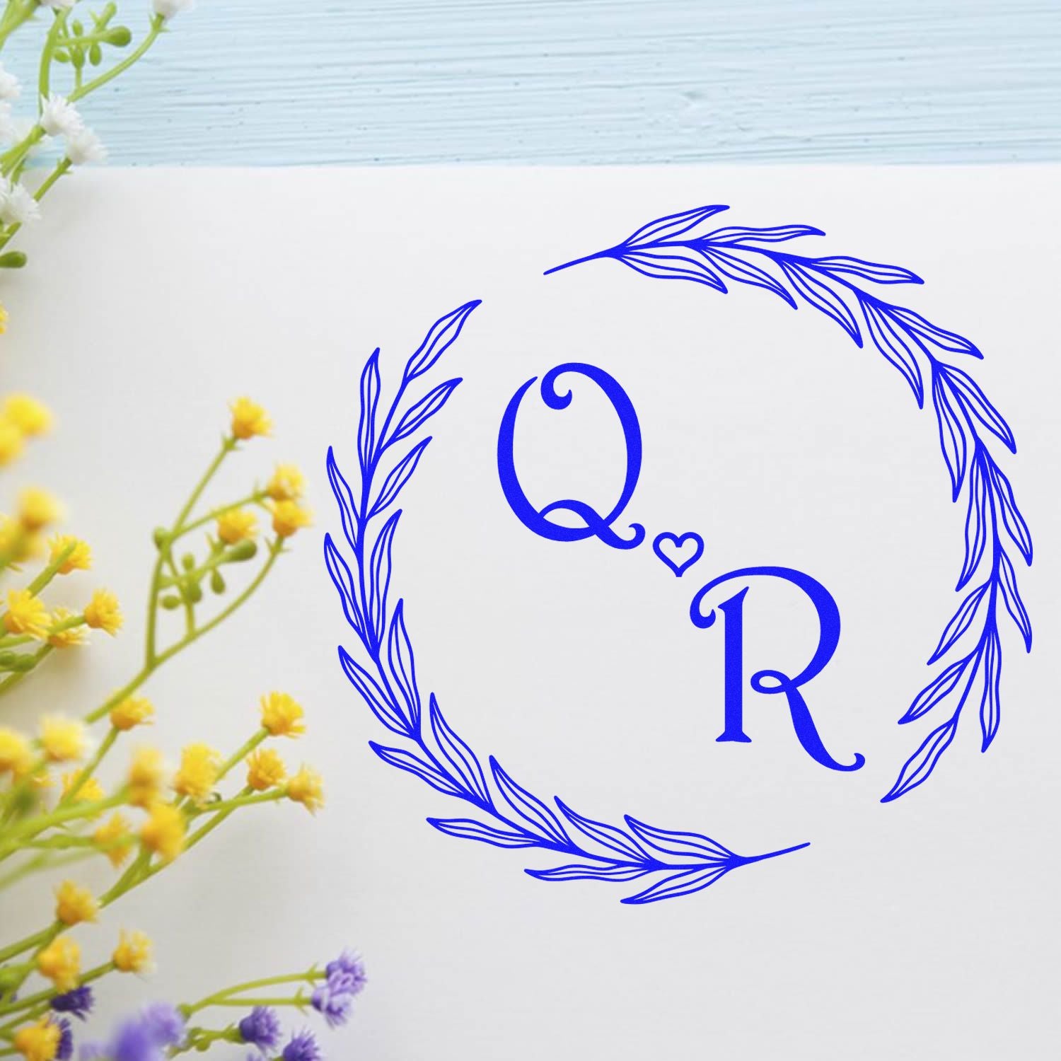 Blue Q?R monogram stamp and seal in a leafy circular wreath on white paper, with yellow and purple flowers at the edges.