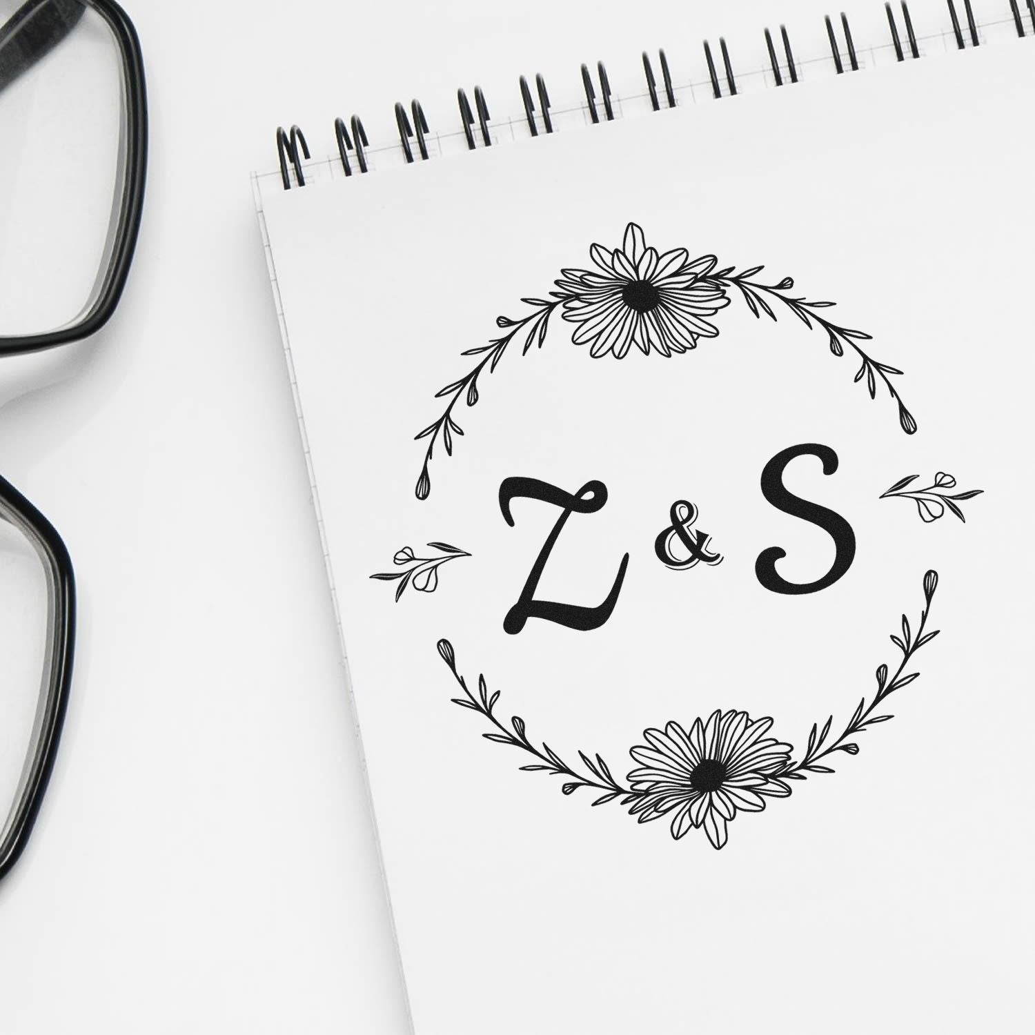 Black floral Z & S stamp and seal design on a white spiral notebook, with delicate wreath details and eyeglasses.