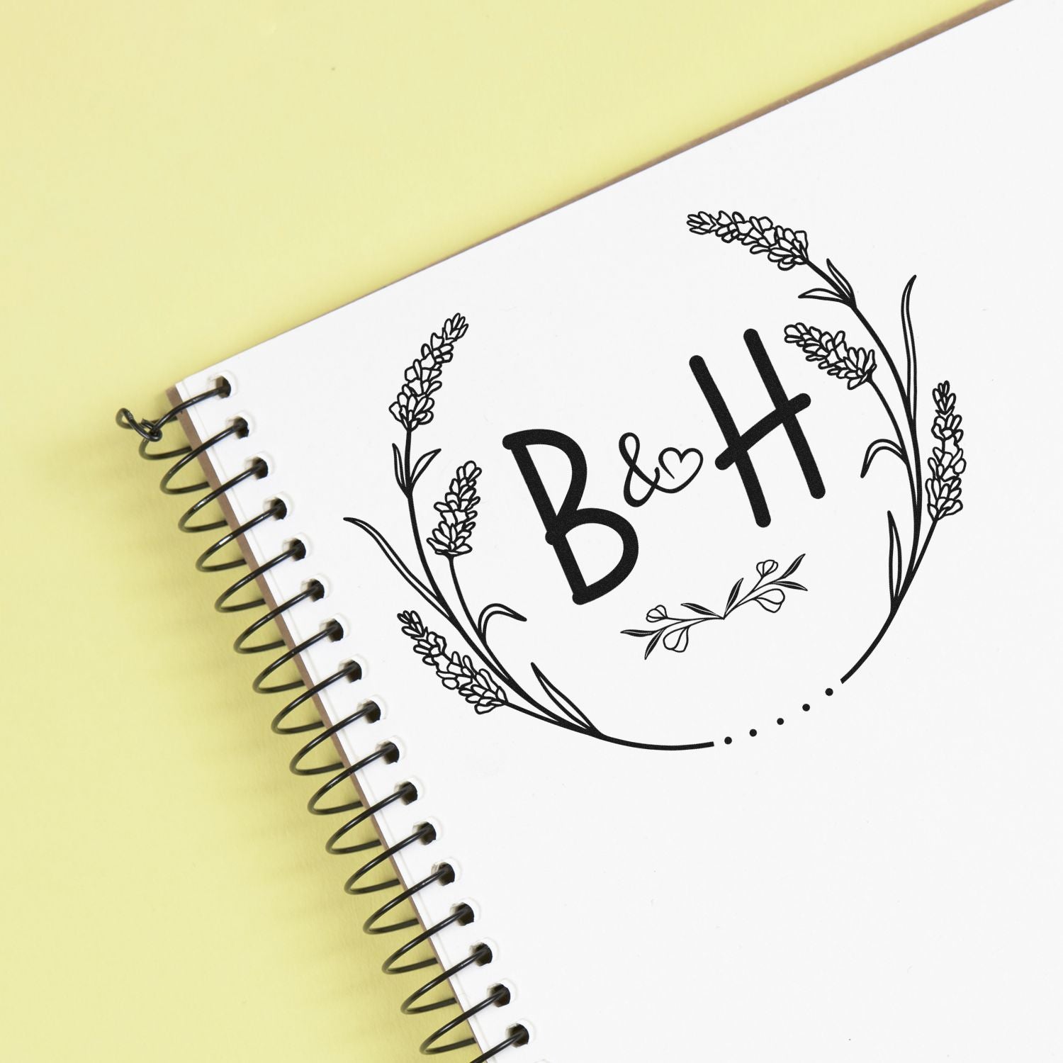 Black floral custom stamp seal with B&H on a white spiral notebook, set against a pale yellow background.