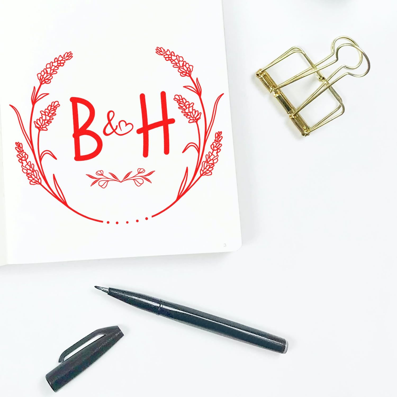 Red custom stamp and seal design with B&H monogram, floral wreath, gold clip, and black pen on white paper.