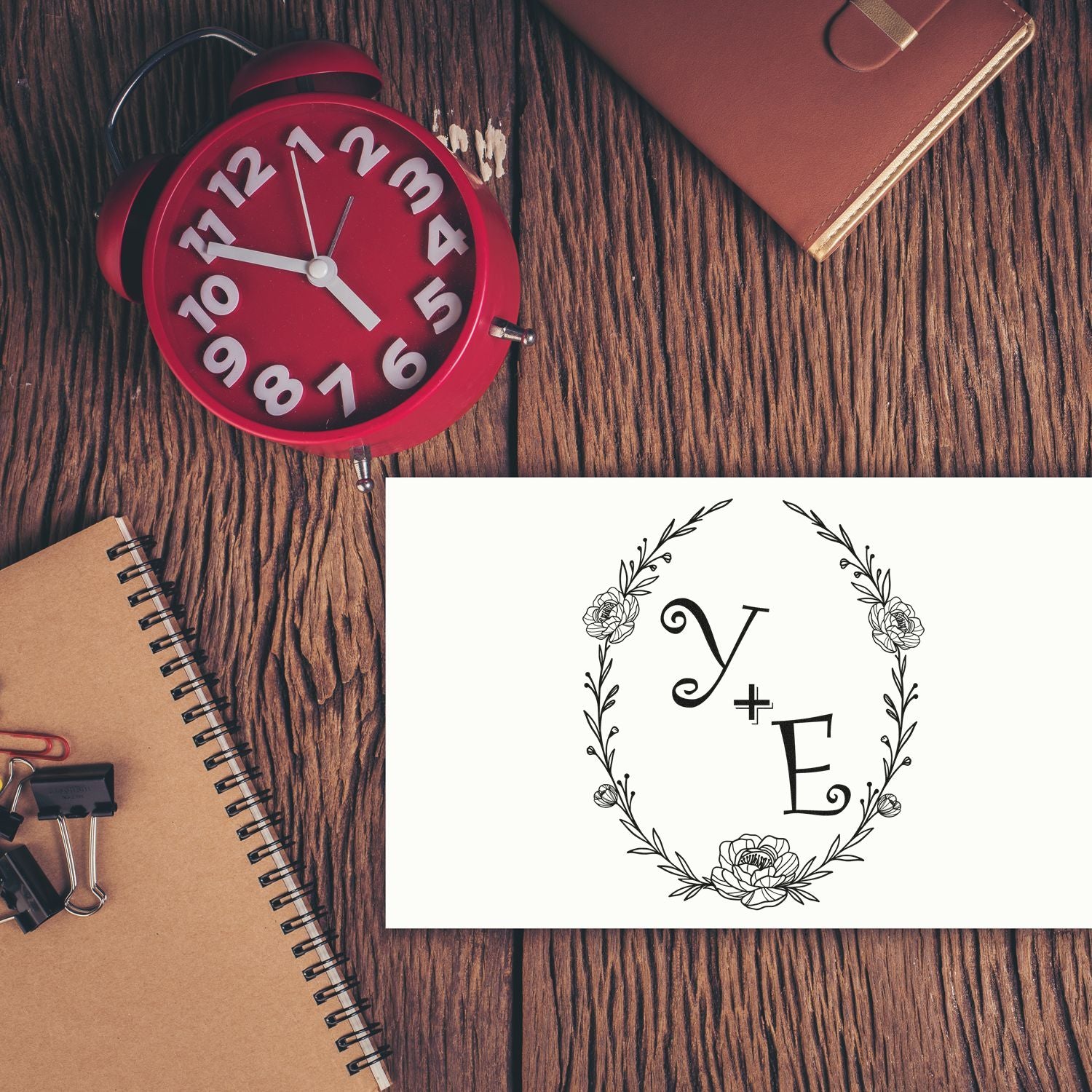 Red alarm clock beside notebooks and a white floral Y+E monogram stamp seal on a dark wood desk.