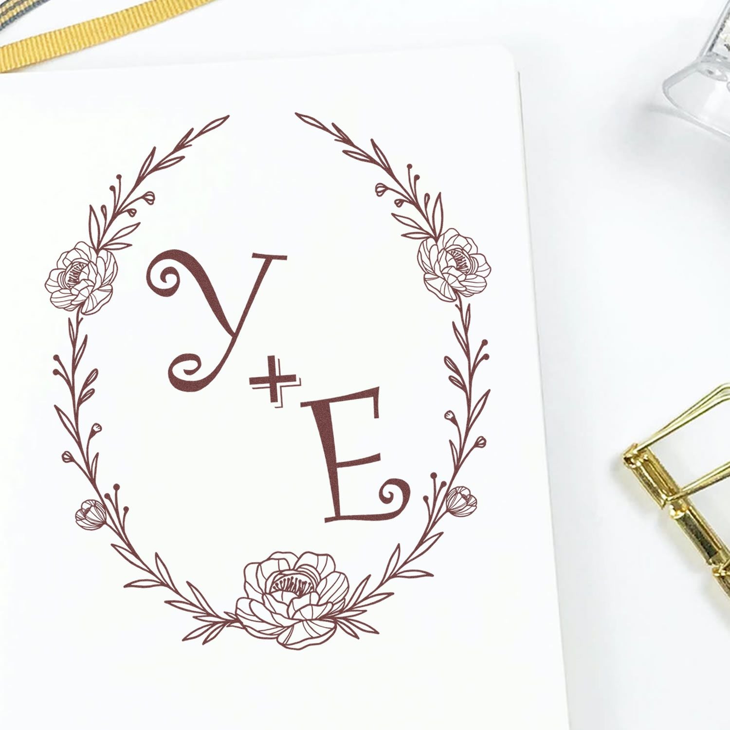 Brown floral monogram wax seal stamp with Y+E initials, wreath and flowers on white card, with gold stamp tool nearby