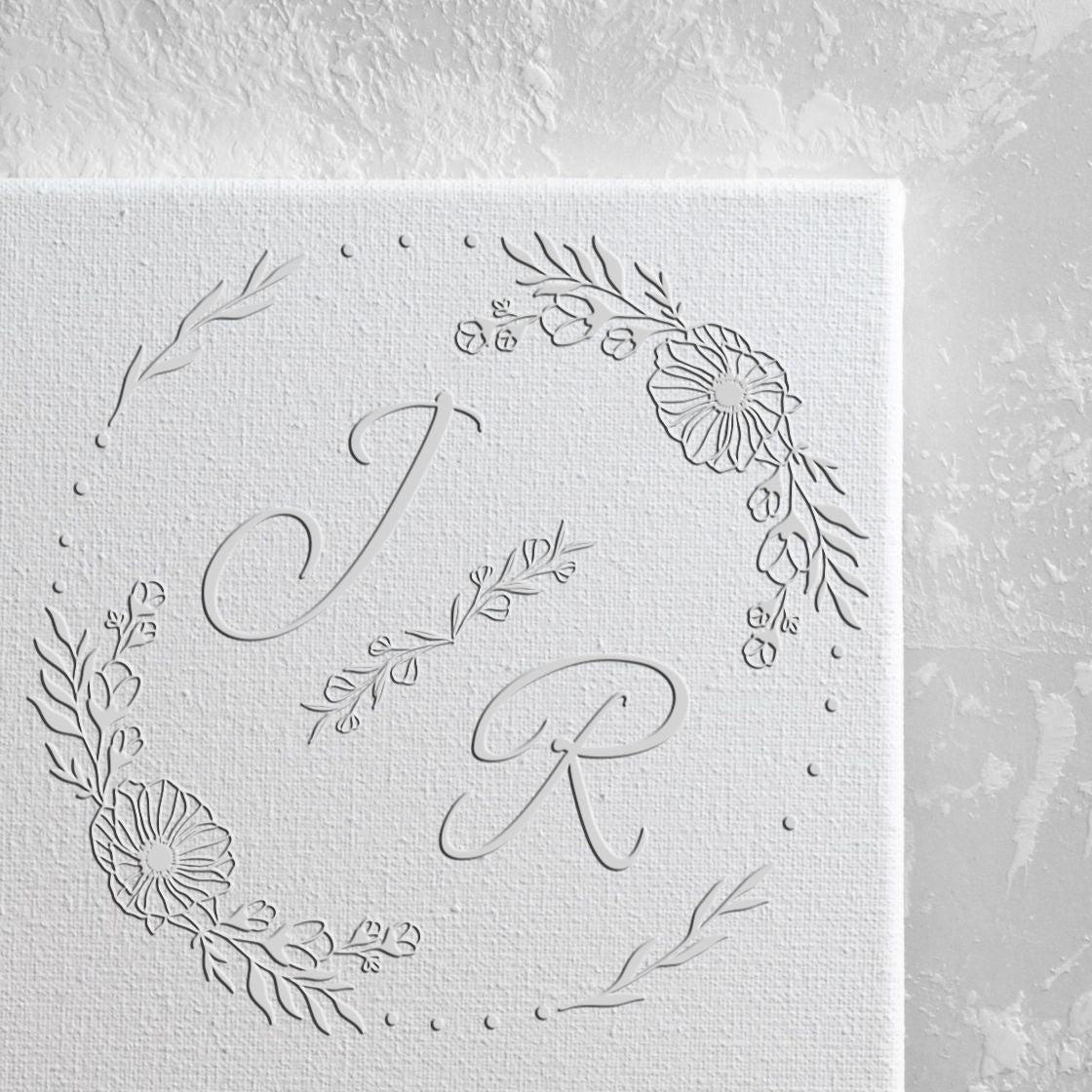 White custom stamp and seal with embossed J R monogram and delicate floral wreath on textured white paper