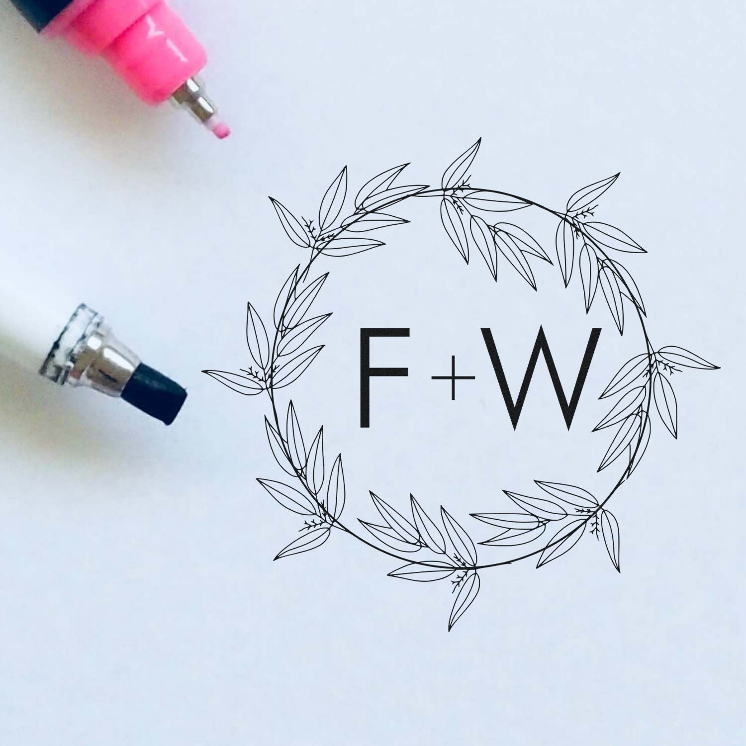 Pink and navy stamp tools beside a black leafy wreath seal with F+W monogram on white paper.
