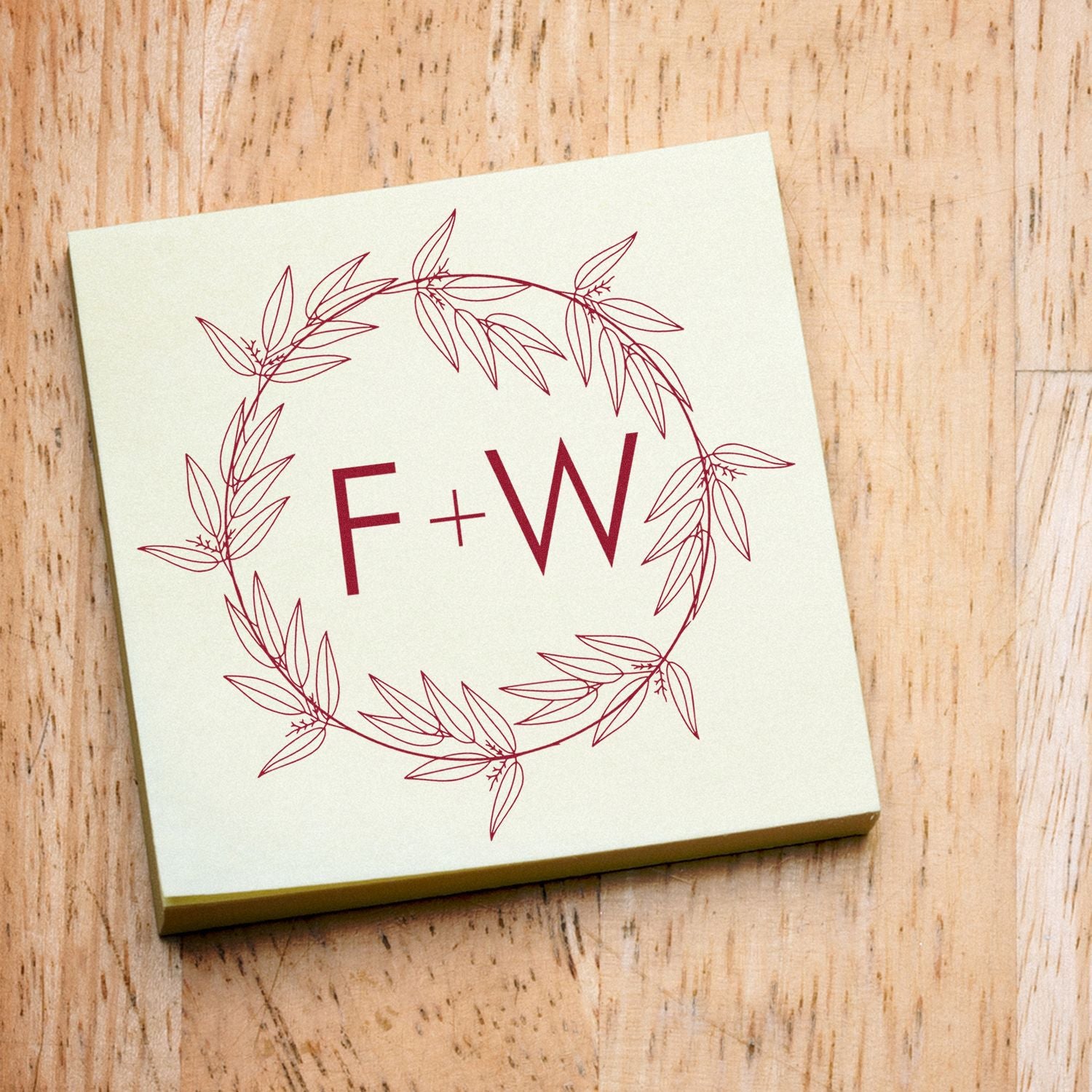 Cream square custom stamp seal with burgundy F+W monogram inside a leafy wreath on a light wood surface.