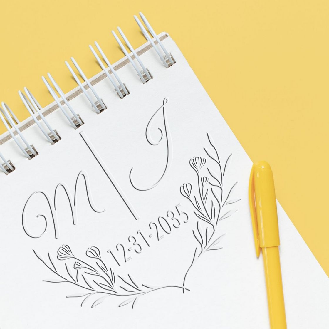 White spiral notebook showing a monogram stamp and floral seal design, with a yellow pen on a yellow background.