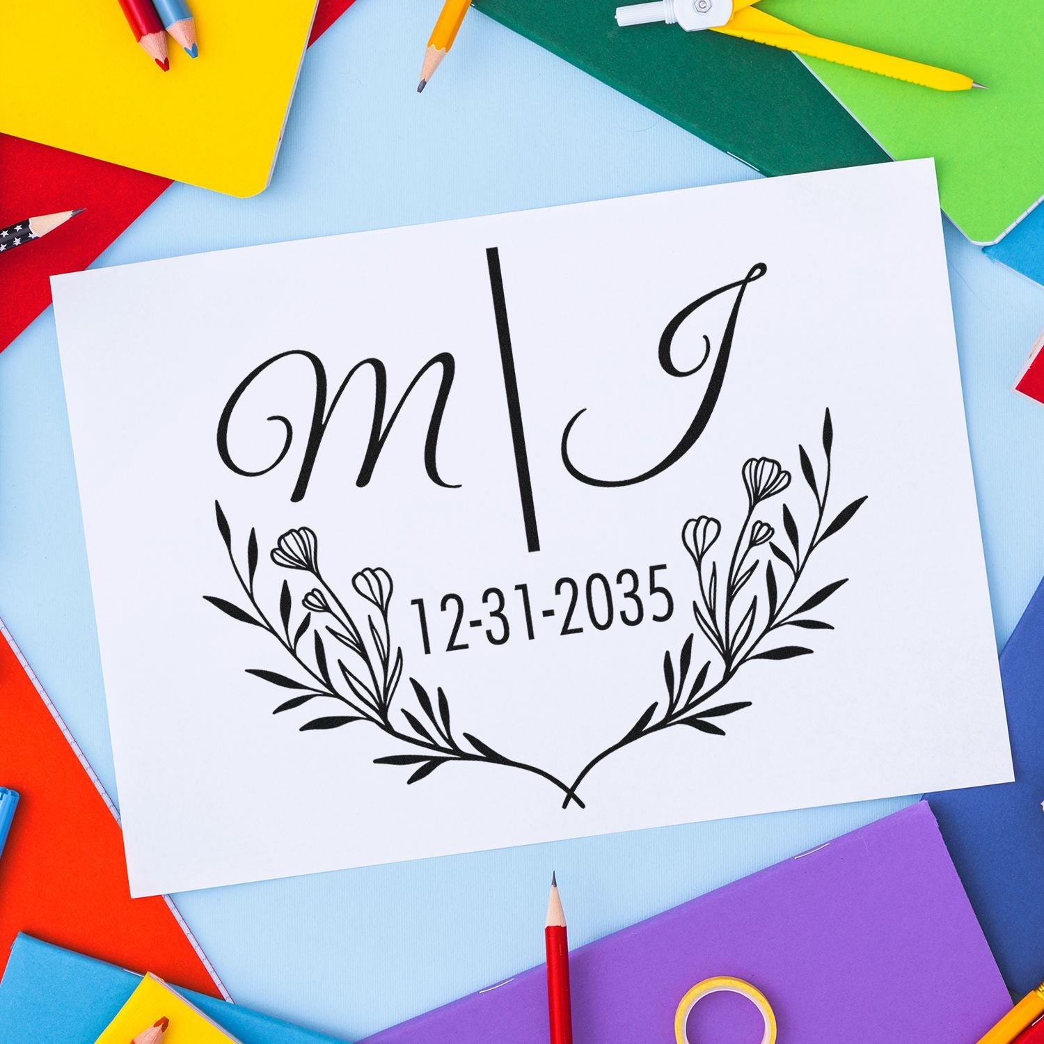 Custom stamp seal with M and J, floral wreath and date 12-31-2035 on white card, framed by colorful pencils and notebooks.