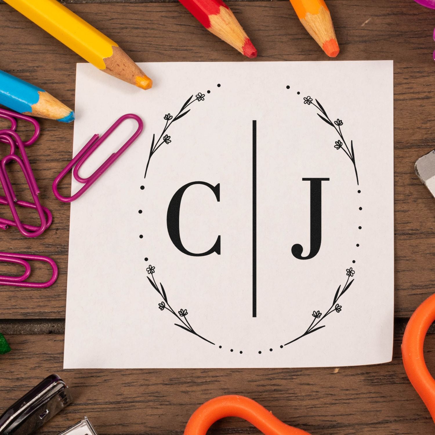 Black CJ stamp and seal monogram on white card, framed by floral wreath, with colorful pencils and paper clips.