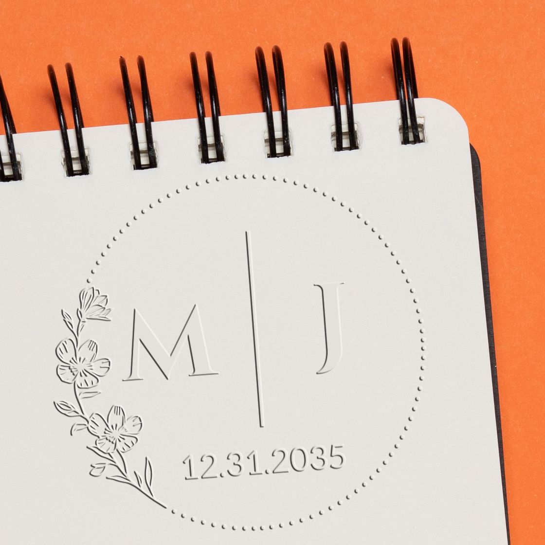Embossed custom stamp seal on white notebook page, with floral monogram M J and date 12.31.2035 on orange background.