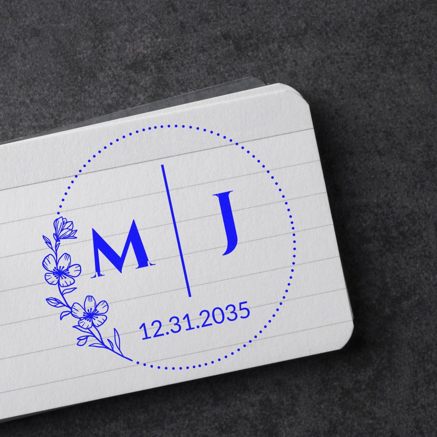 Blue floral monogram stamp seal with M and J, dotted circle, and date 12.31.2035 on white card over dark background.