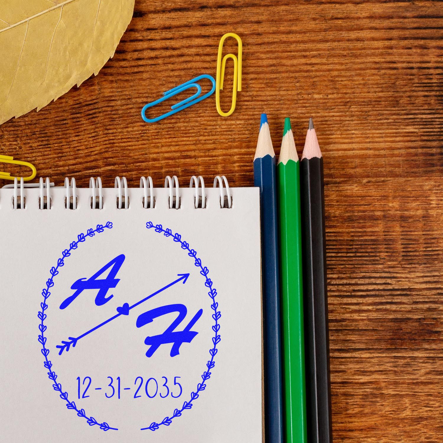 Blue custom stamp and seal design on a spiral notebook, with date 12-31-2035, beside colored pencils and paper clips on wood.