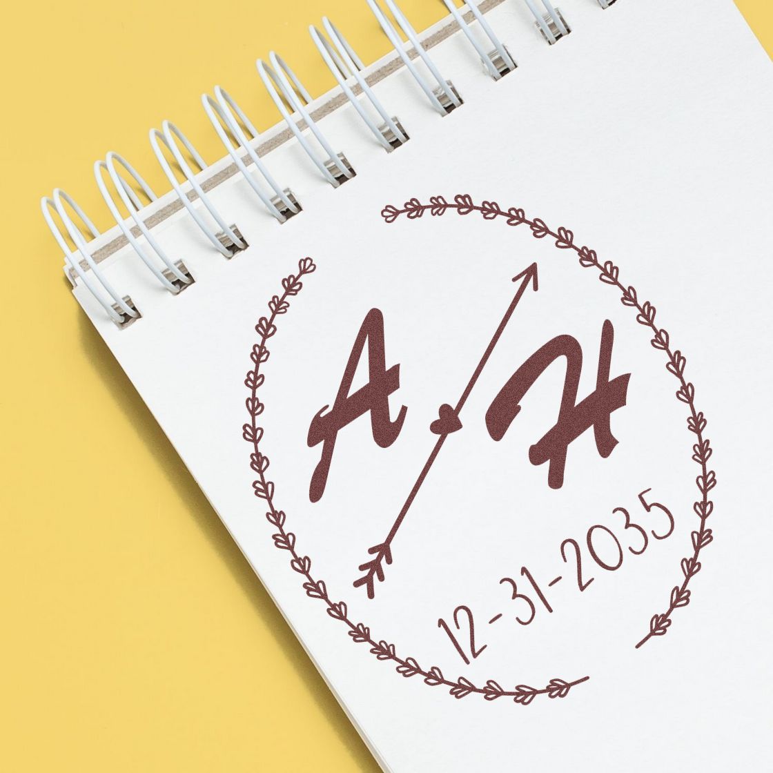 Brown custom stamp seal design on a white spiral notebook page, with leafy wreath, arrow, initials, and date 12-31-2035 on yellow background.