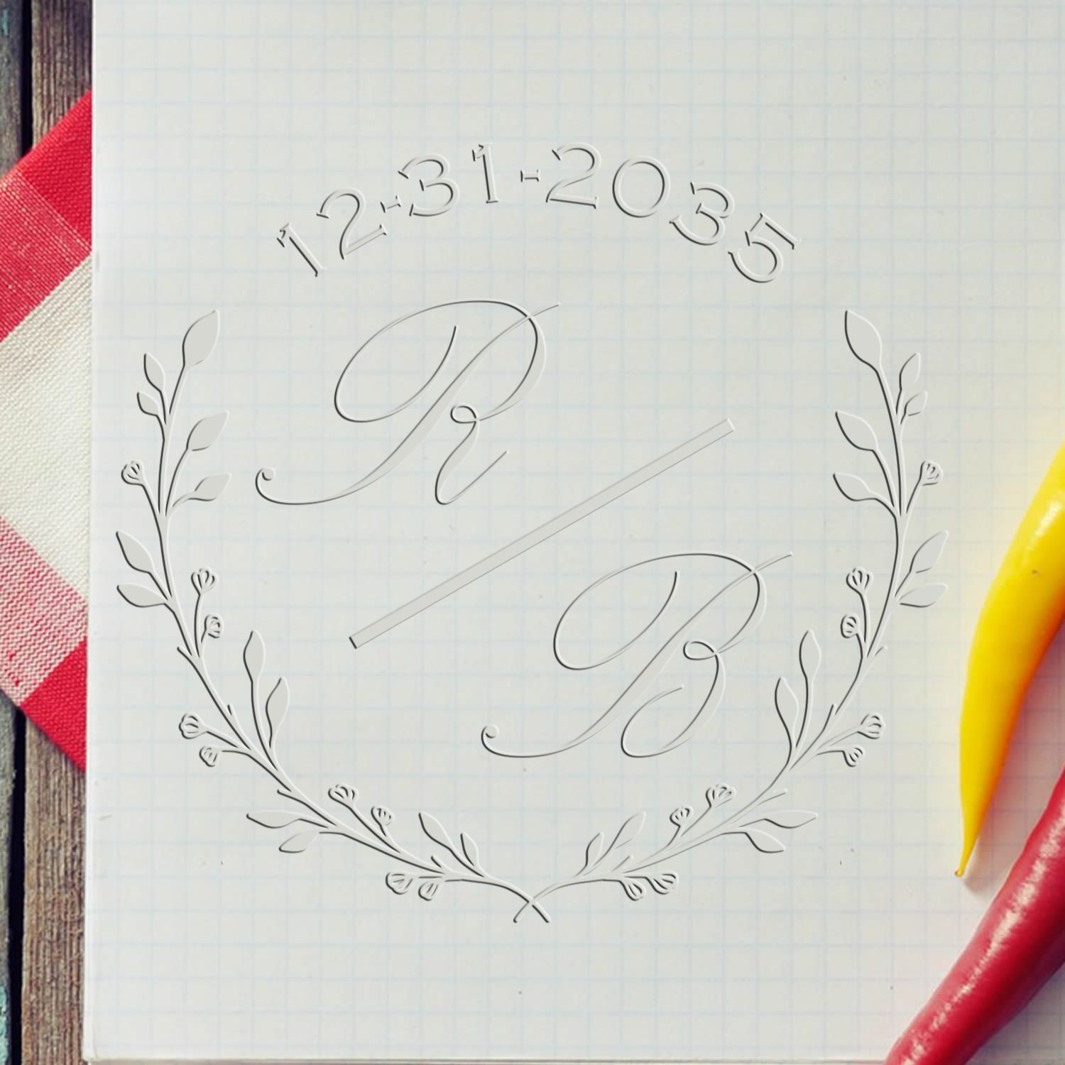 Clear acrylic stamp seal with floral wreath, script monogram and arched numbers on white grid paper, beside red and yellow pens.