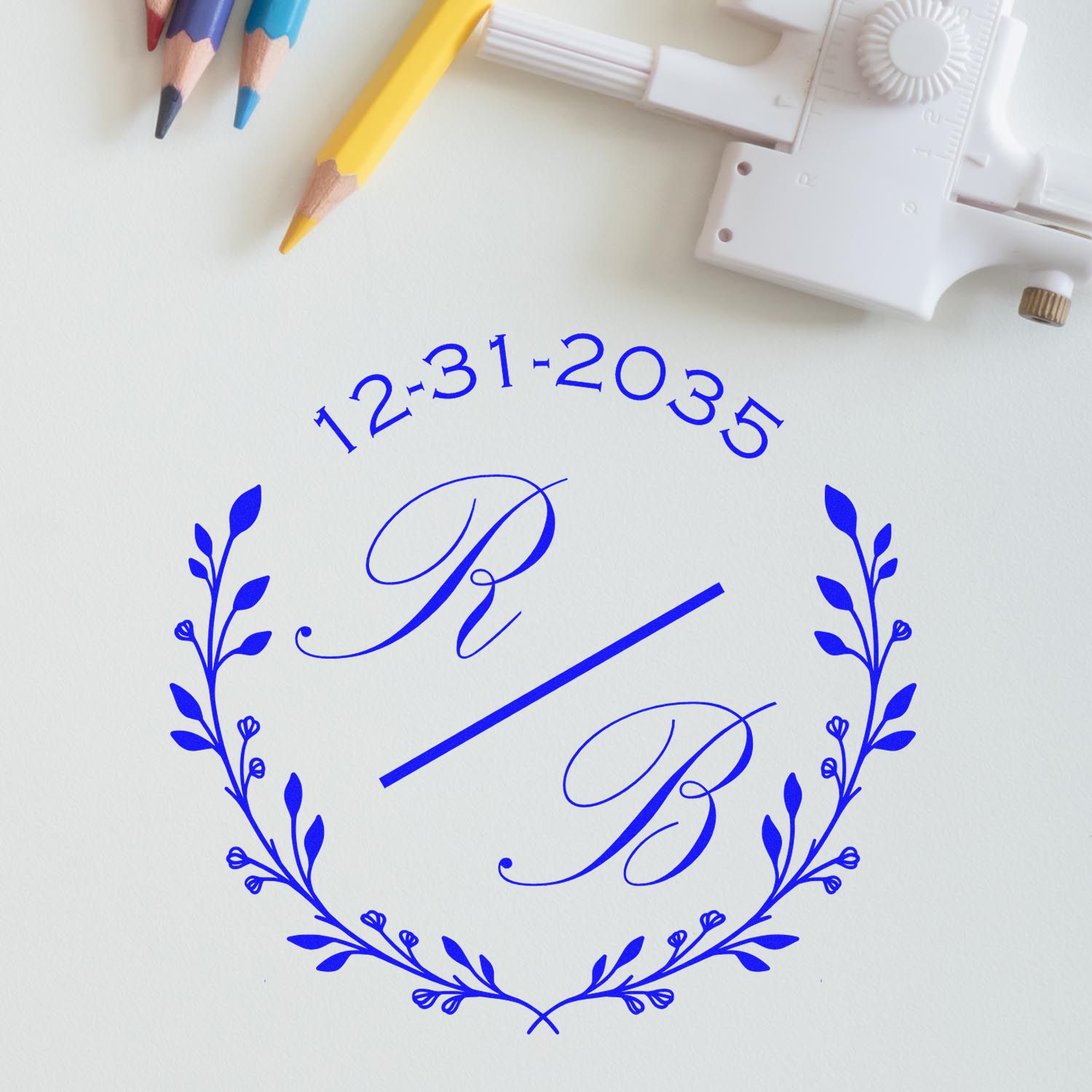 Blue custom stamp and seal with floral wreath, monogram P and B, and date 12-31-2035 on white paper beside colored pencils.