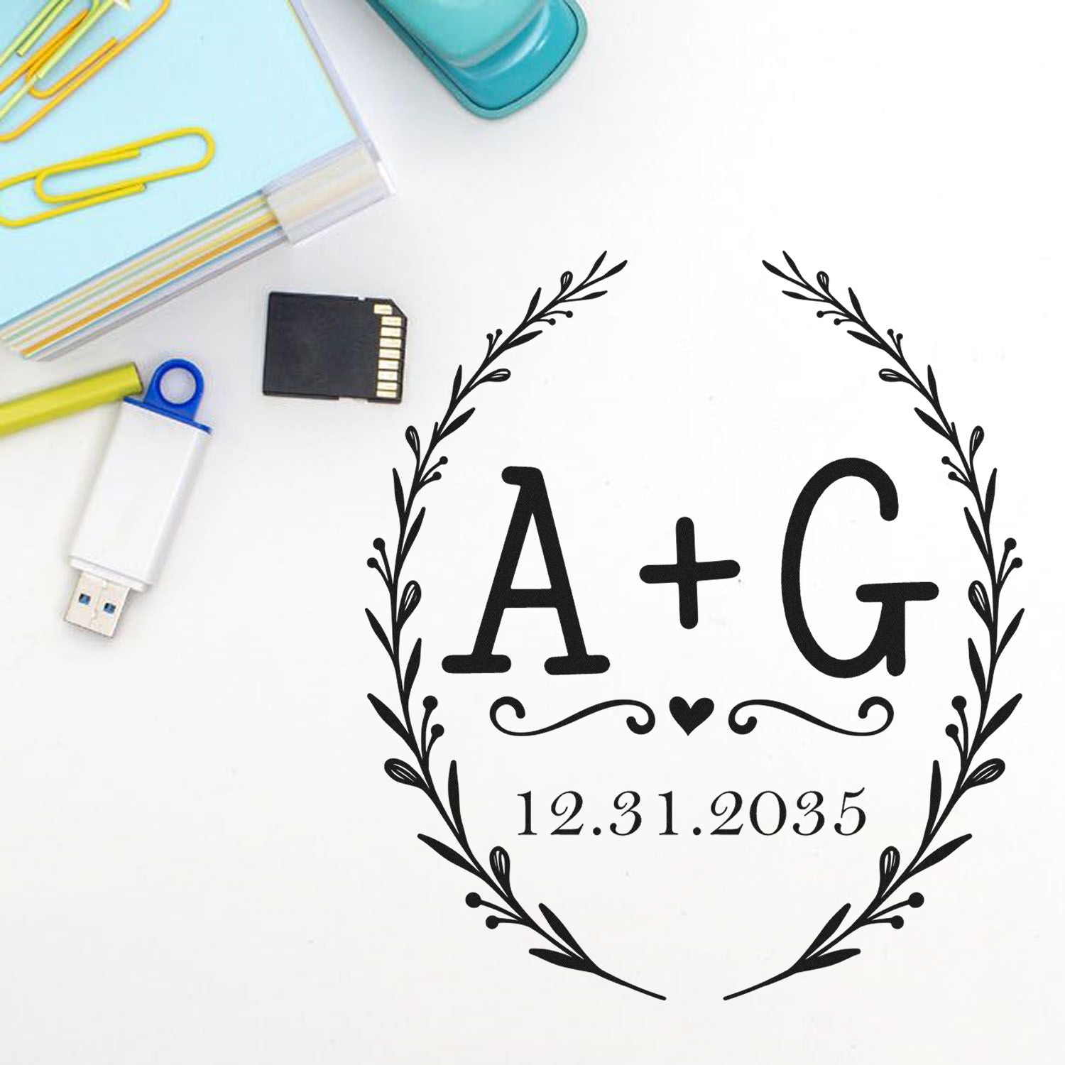 Black custom stamp and seal with laurel wreath, A+G monogram and 12.31.2035 date on white, beside pastel office supplies.