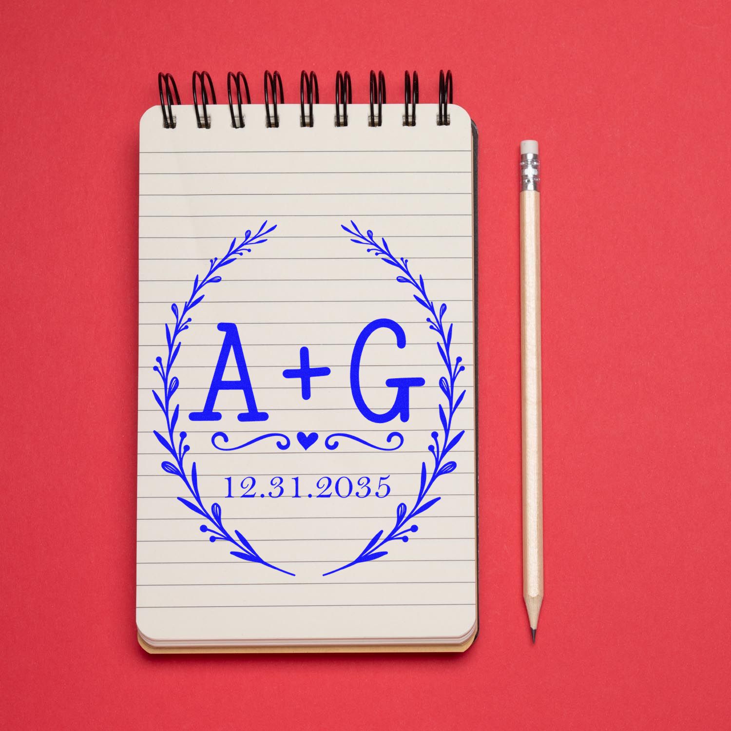 Custom blue A+G wedding seal stamp with wreath and 12.31.2035 on lined notebook, beside pencil on red background.