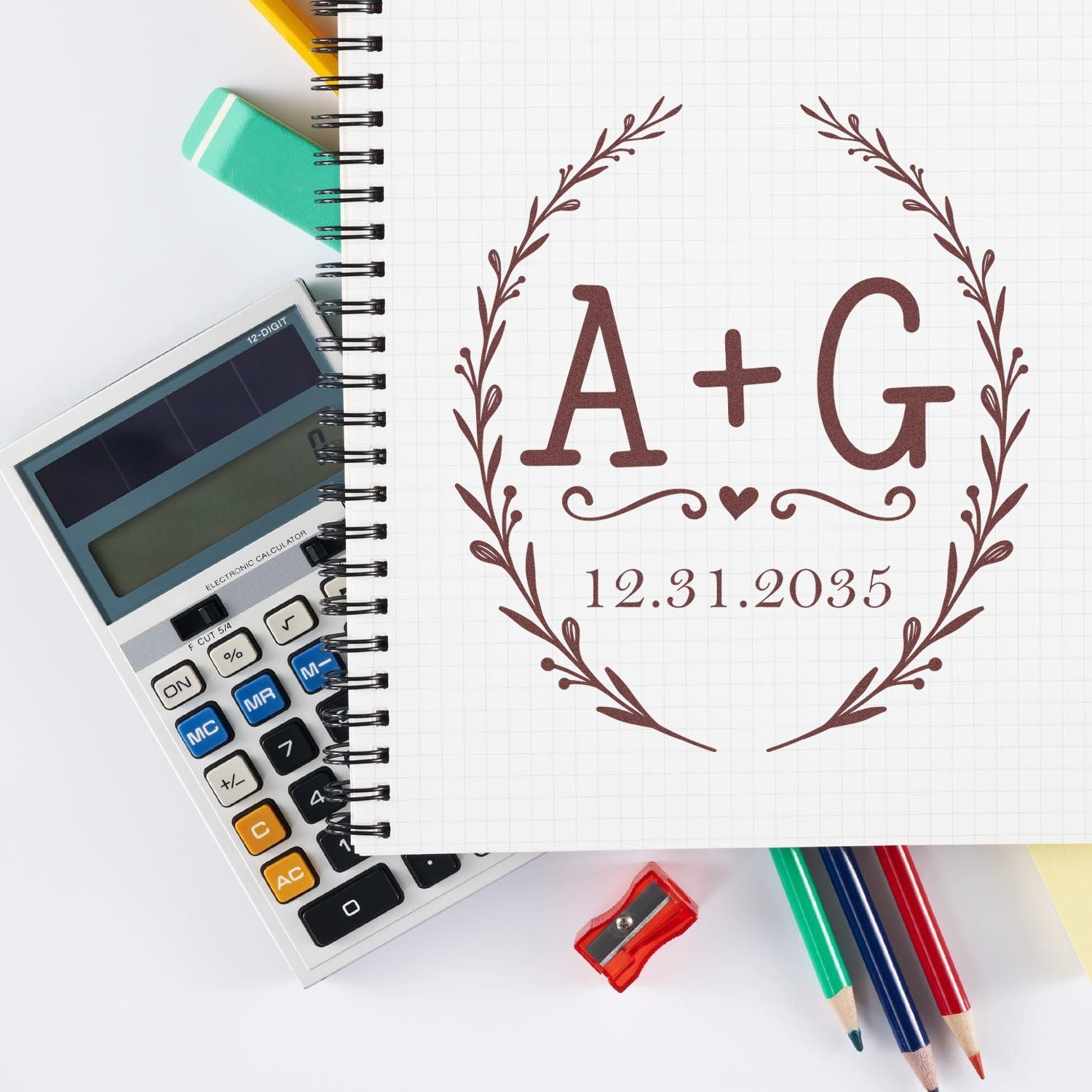 Brown A+G custom seal stamp with laurel wreath and date on grid notebook, beside calculator, pencils, and erasers.