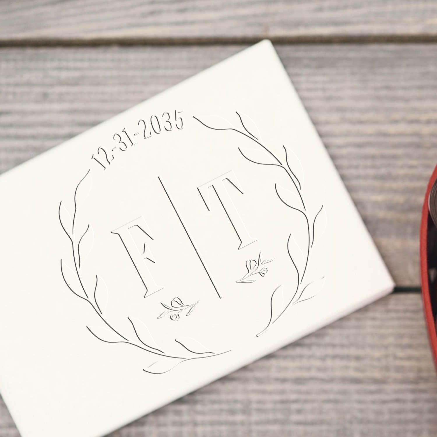 White custom stamp and seal with embossed FT monogram, 12-31-2035 date, and leafy wreath on gray wood background.
