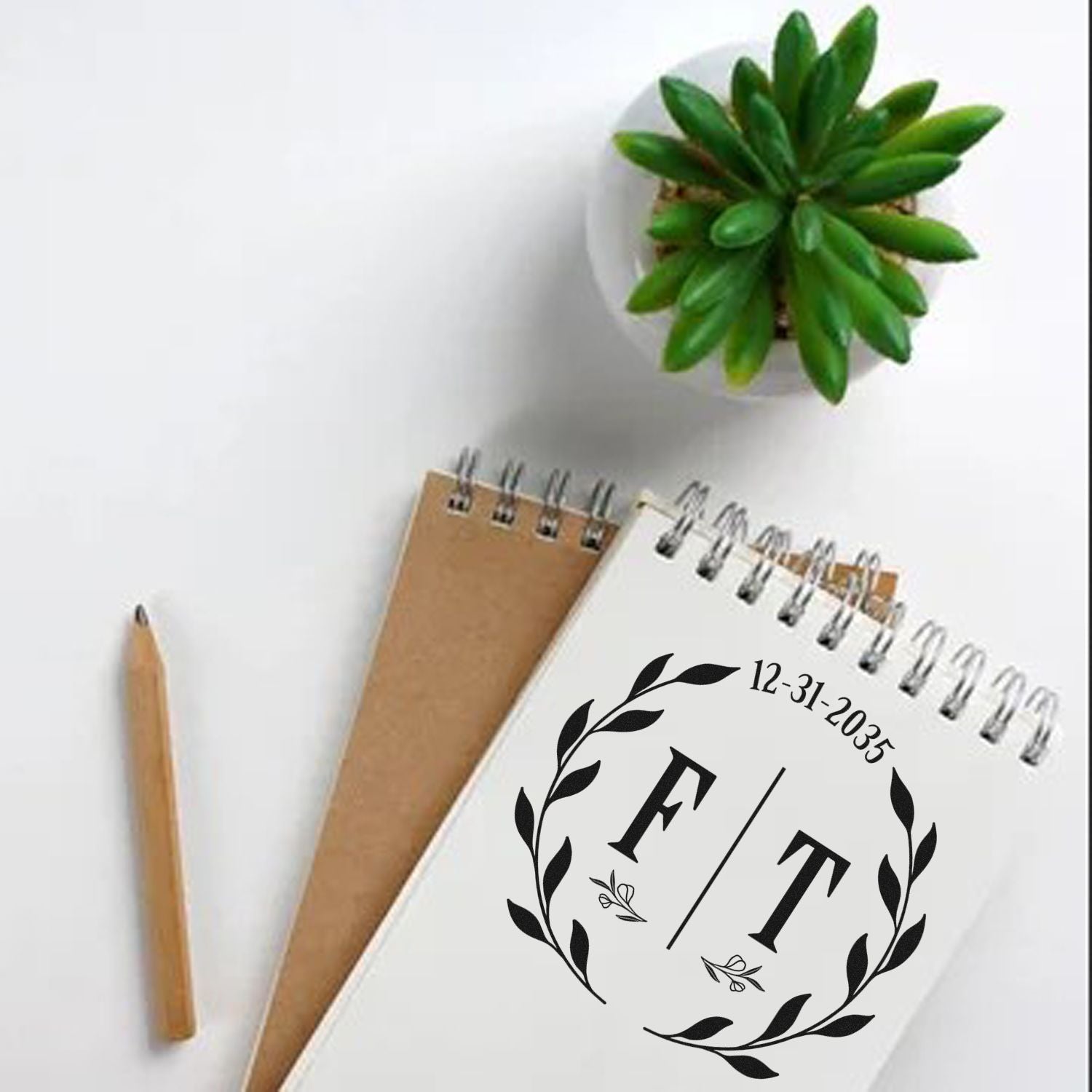 Custom black laurel monogram stamp and seal on white spiral notebook, with date, kraft notebook, pencil, and green succulent.
