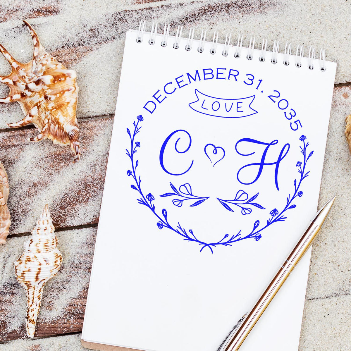 Blue custom stamp and seal with monogram, floral wreath, LOVE banner, and December 31, 2035 on white notebook beside seashells.