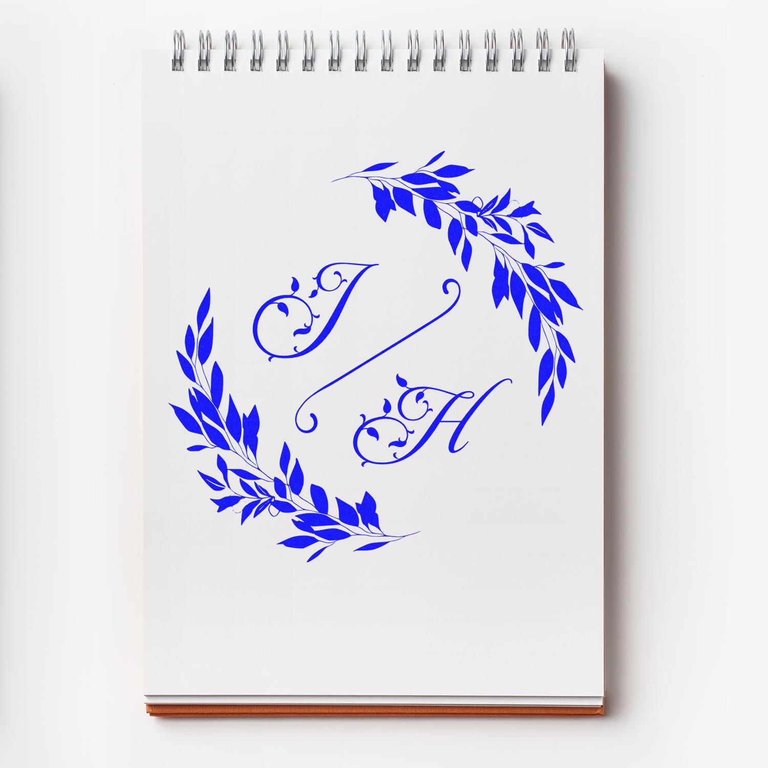 Blue custom stamp and seal monogram J H with leafy wreath on a white spiral notebook