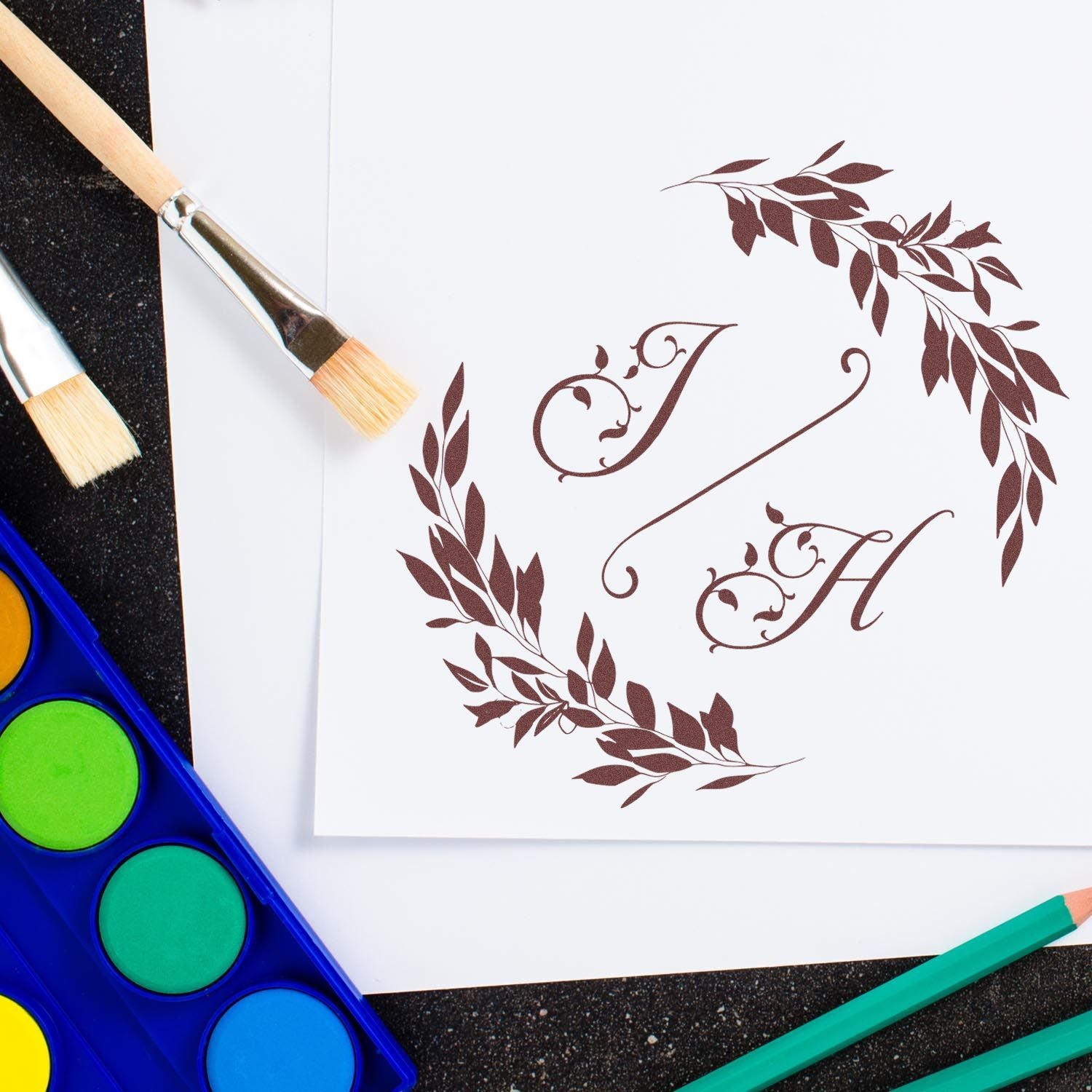 Brown floral monogram stamp and seal on white paper, surrounded by paintbrushes, watercolor palette, and green pencils.