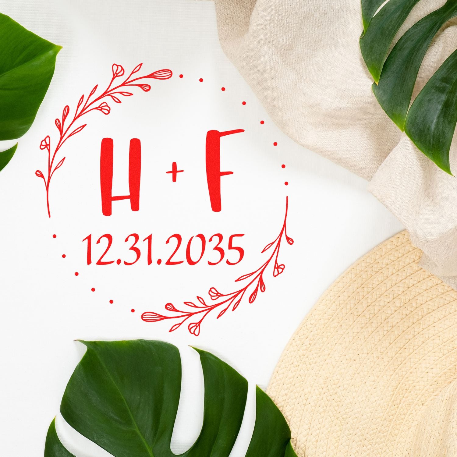 Red wedding stamp seal with H+F and 12.31.2035, floral wreath, on white with green leaves, beige fabric and straw hat.