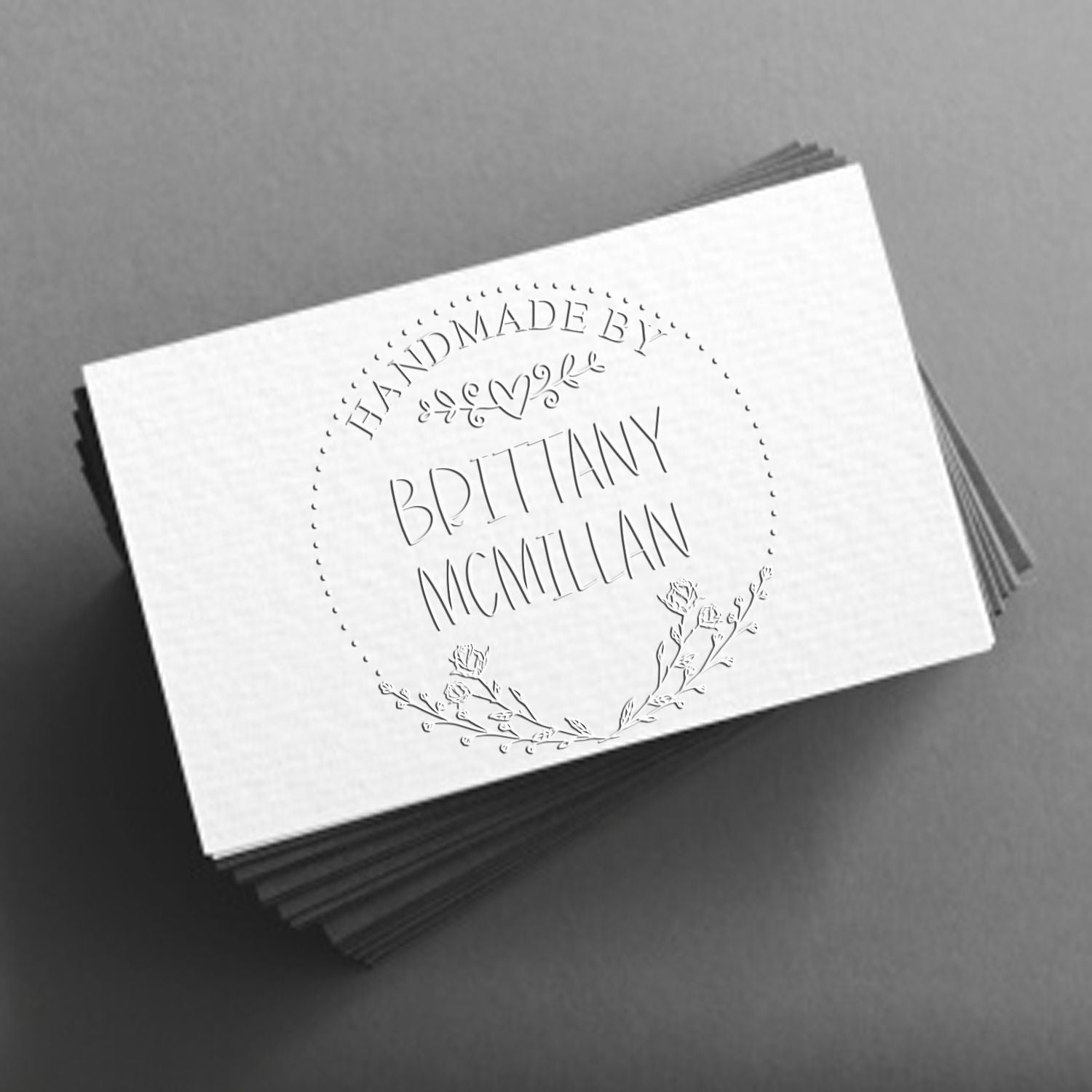 Stack of white custom stamp and seal cards with embossed floral wreath and Handmade by Brittany McMillan on a gray background