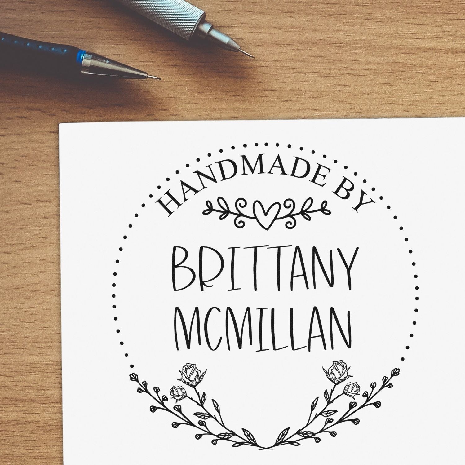 Custom black handmade-by stamp seal for Brittany McMillan, with floral wreath on white paper and pens on wood desk.