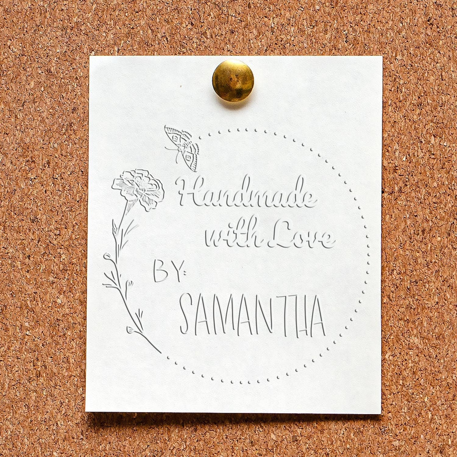 White embossed stamp seal sample with gold pin, floral butterfly detail, and handwritten wedding text on cork board.