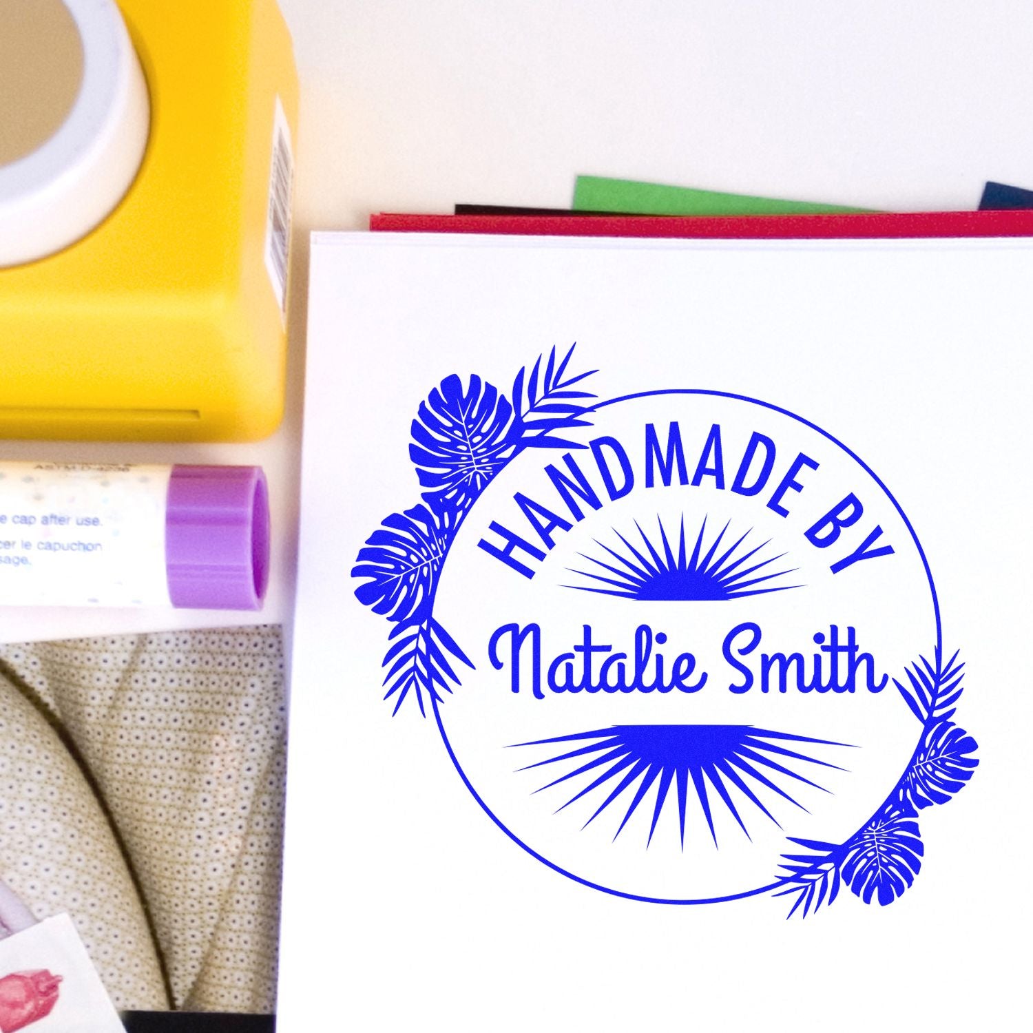 Custom blue handmade seal stamp for Natalie Smith with tropical leaves on white paper, beside yellow and purple stamp supplies.