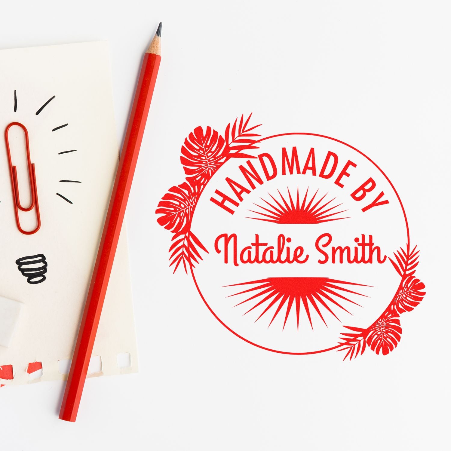 Red custom stamp and seal reading Natalie Smith, with tropical leaves, beside a red pencil and paper clips.