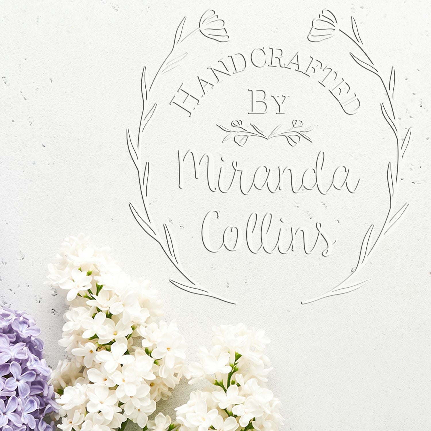 Embossed white handcrafted stamp seal with floral wreath, reading Handcrafted by Miranda Collins, beside white and purple flowers.