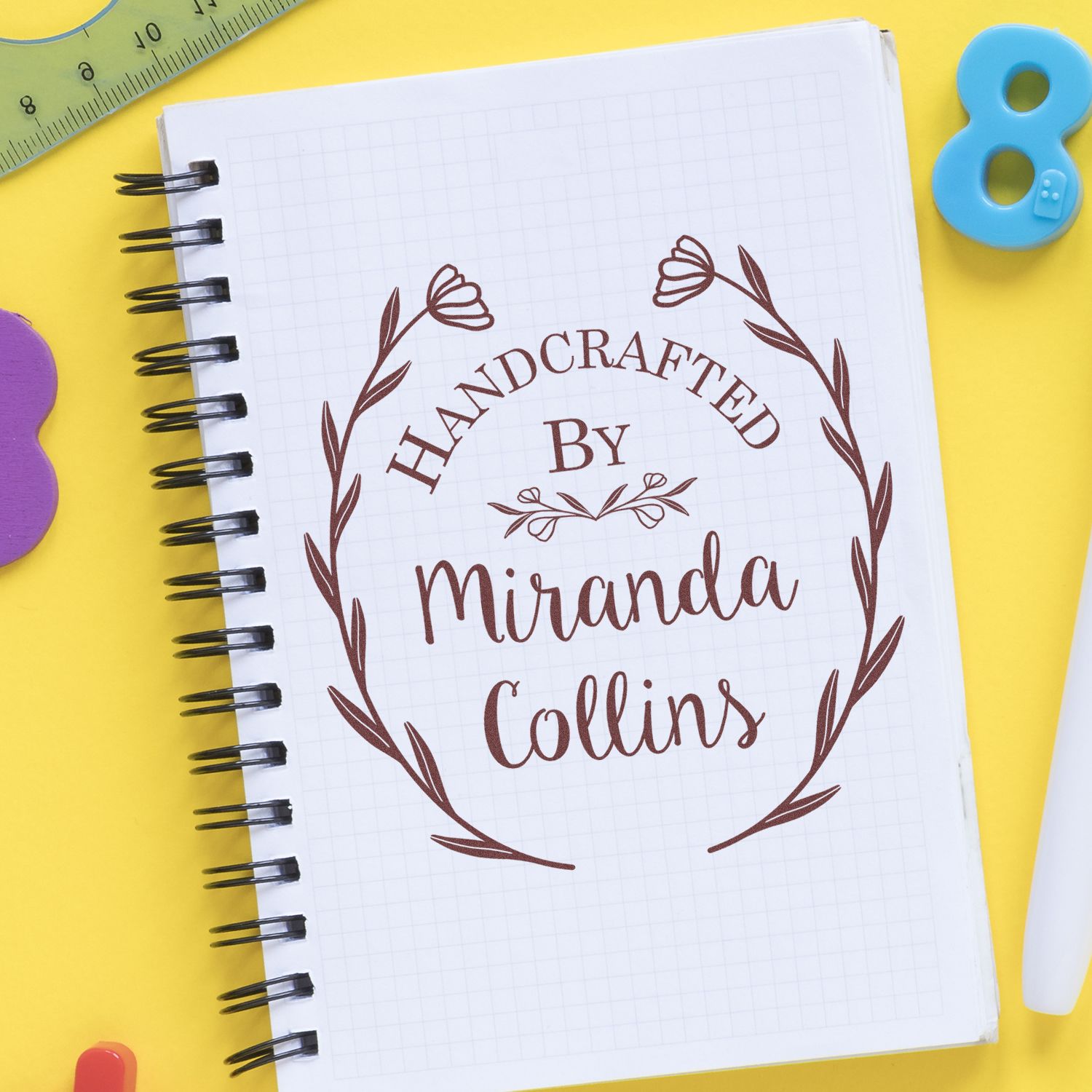 Brown handcrafted stamp and seal with floral wreath on white spiral notebook, on bright yellow background with colorful craft tools.