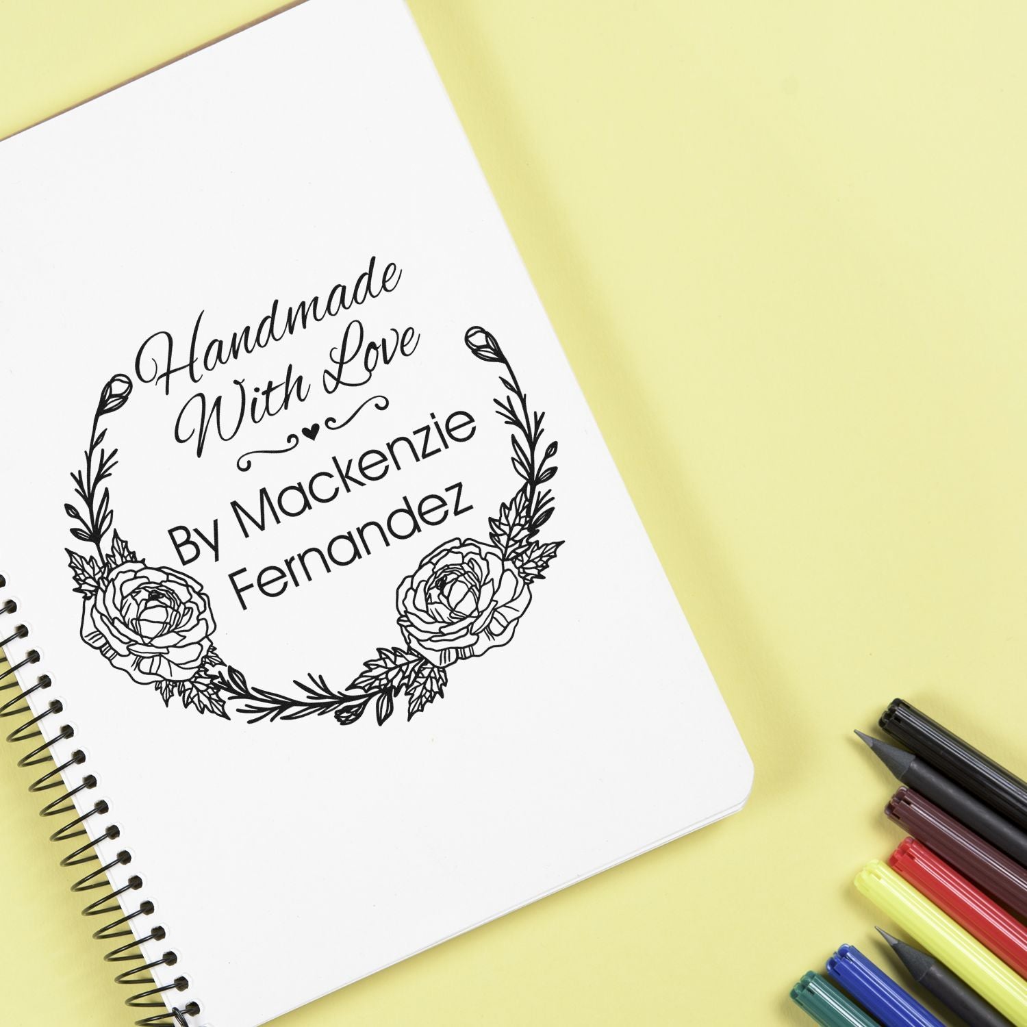 White spiral notebook with a black floral handmade stamp seal reading Handmade With Love, on pale yellow with colorful pens.